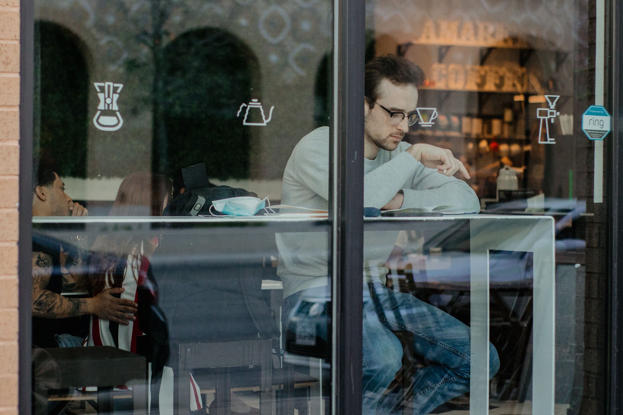 A man with glasses and a beard sitting inside a coffee shop, reading a book. Through the window, a woman with tattoos on her arms is seen, holding a drink and looking outside. The window has icons indicating coffee, tea, and celebration decorations, and a blue sticker that says 'Produced by Ring'.