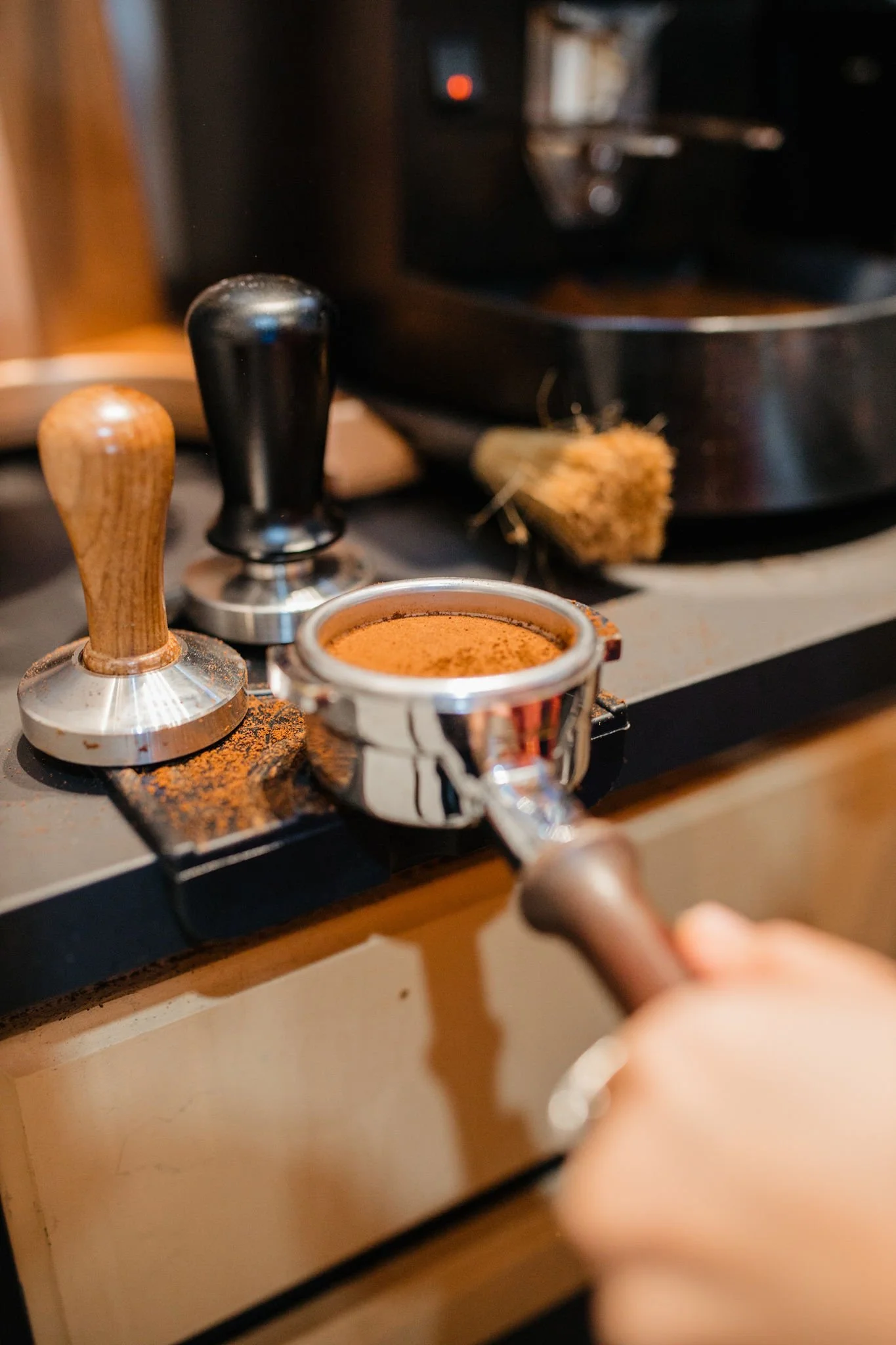 A person is holding a portafilter filled with ground coffee, preparing to make espresso on a stovetop espresso maker, with a moka pot and coffee tamper nearby.