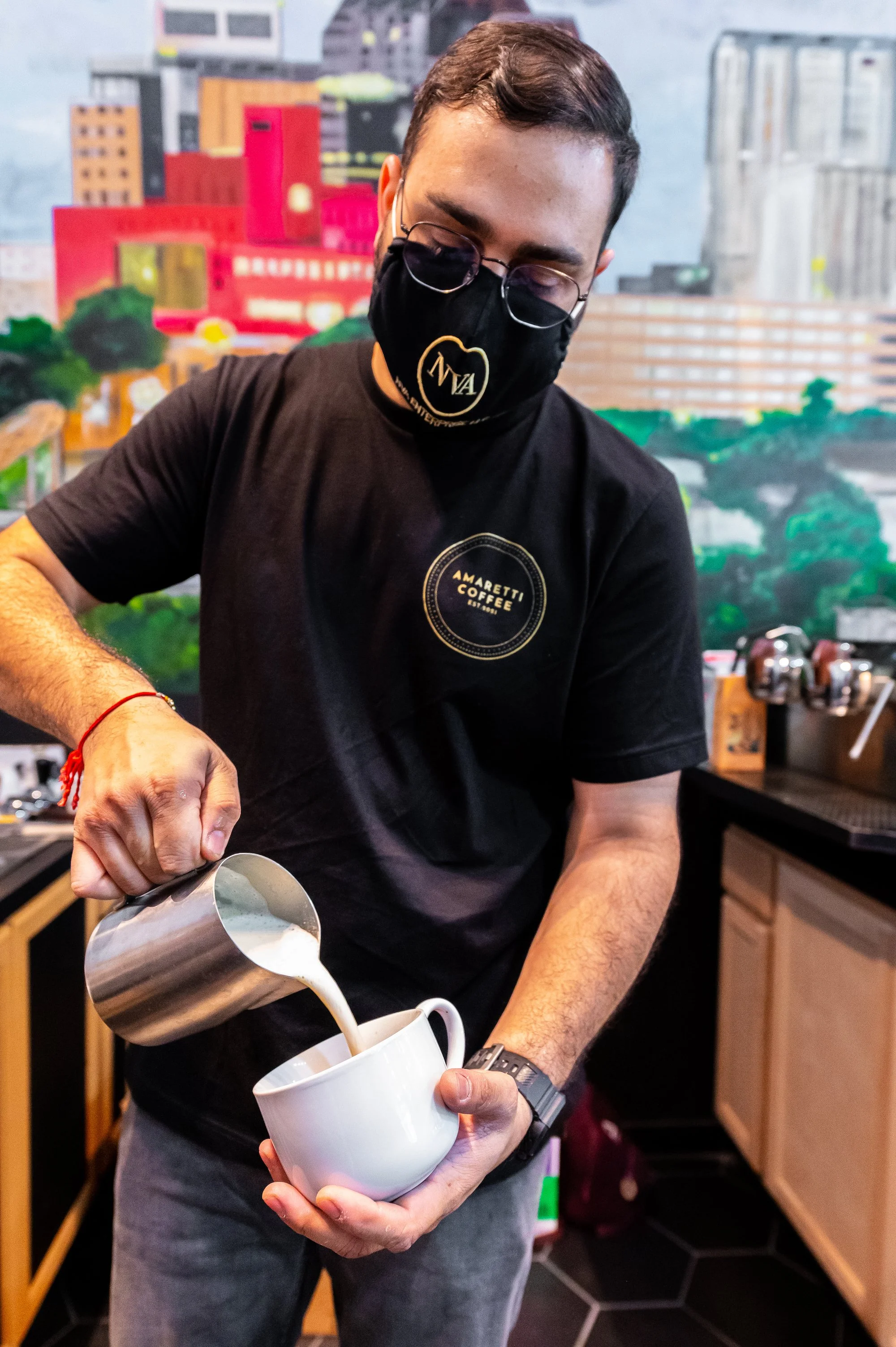 A man with glasses and a black face mask pouring milk from a stainless steel pitcher into a white mug, in a cafe or coffee shop setting.