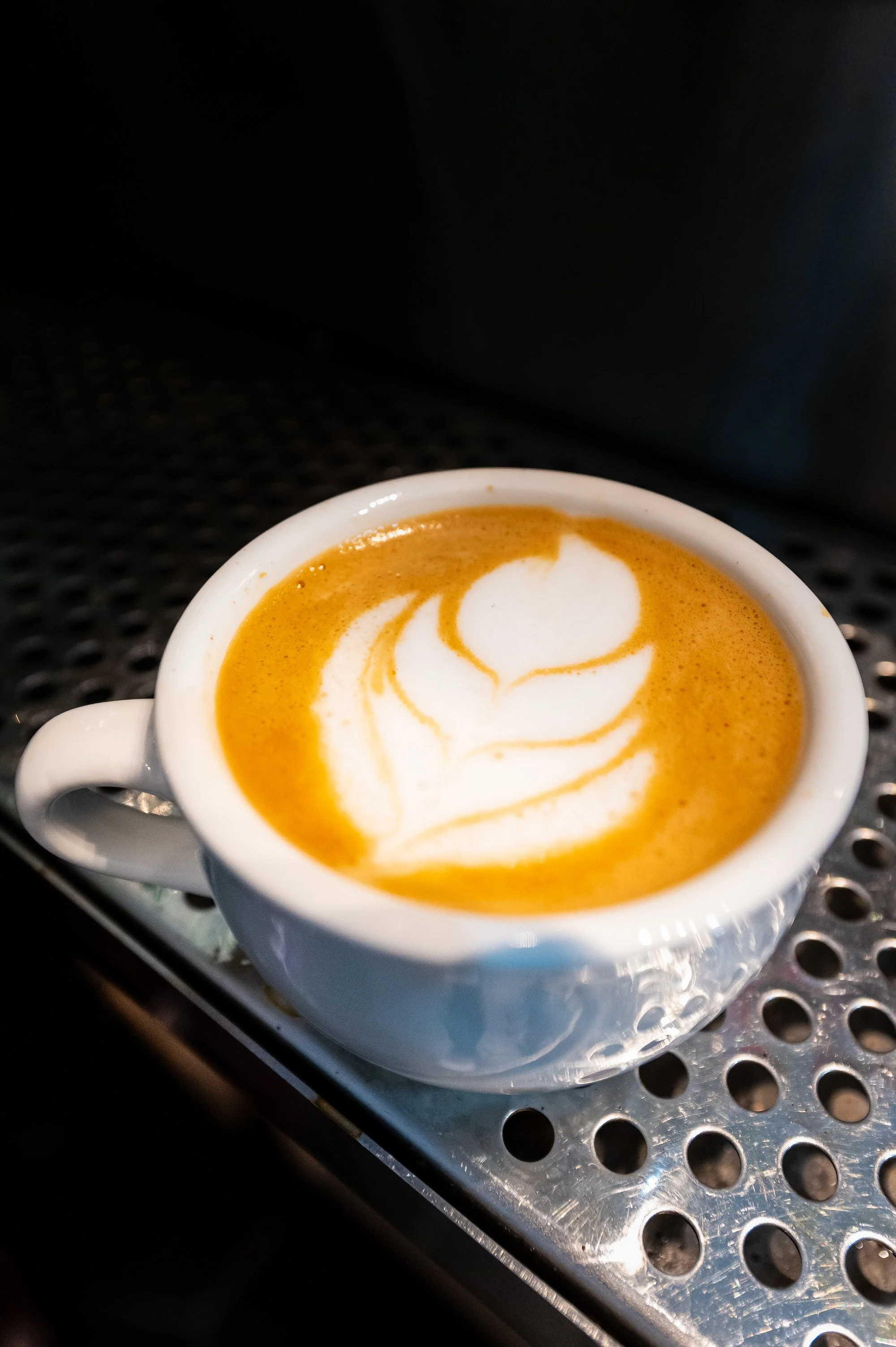 Close-up of a white ceramic mug filled with a coffee beverage topped with latte art, placed on a metal surface with circular holes, with a dark background.