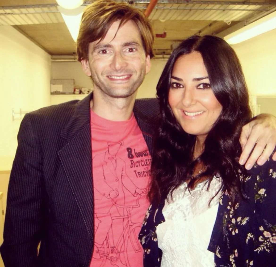 A man and woman smiling and standing close together in an indoor setting, with the man's arm around the woman's shoulder.