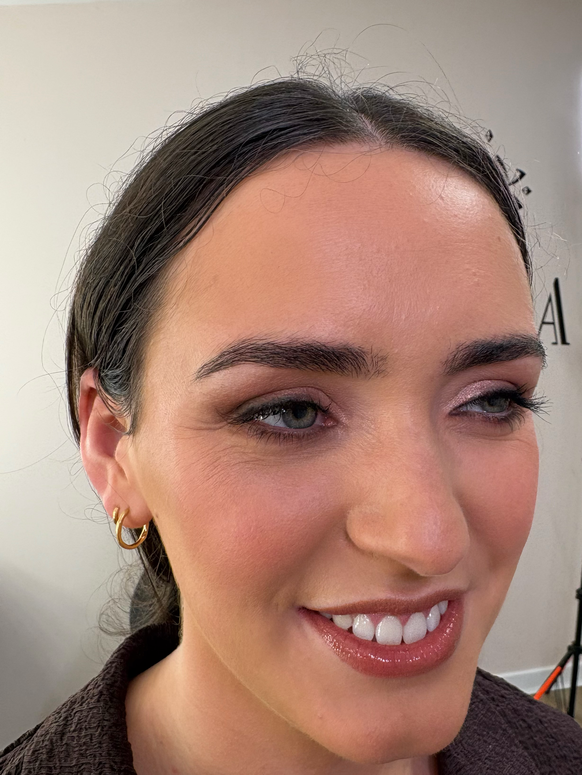 Close-up of a woman smiling with visible teeth, wearing hoop earrings, with dark hair and makeup, in a professional setting.