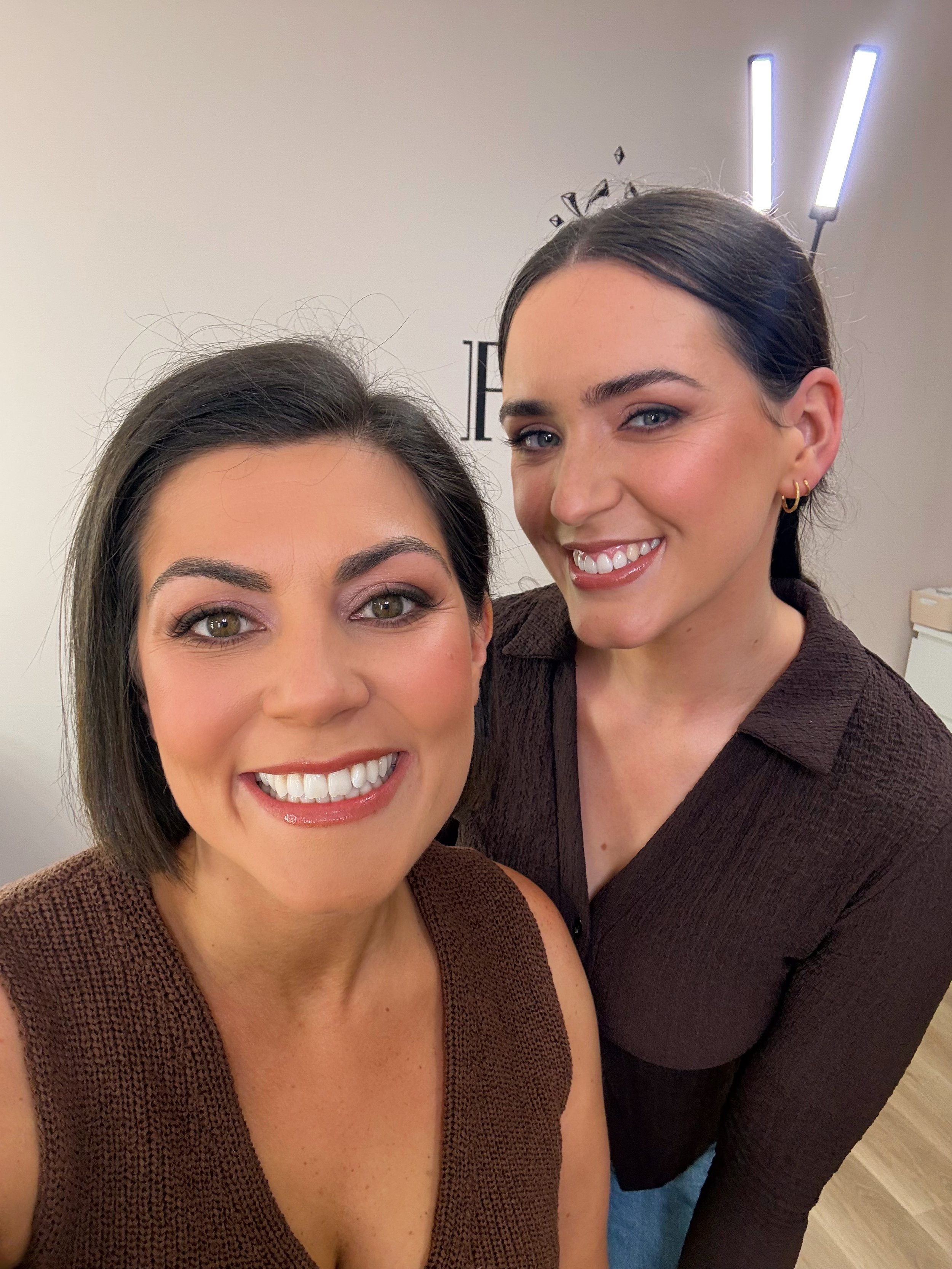 Two women smiling for a selfie, one with dark hair and green eyes, the other with dark hair, blue eyes, and stud earrings, wearing brown tops.