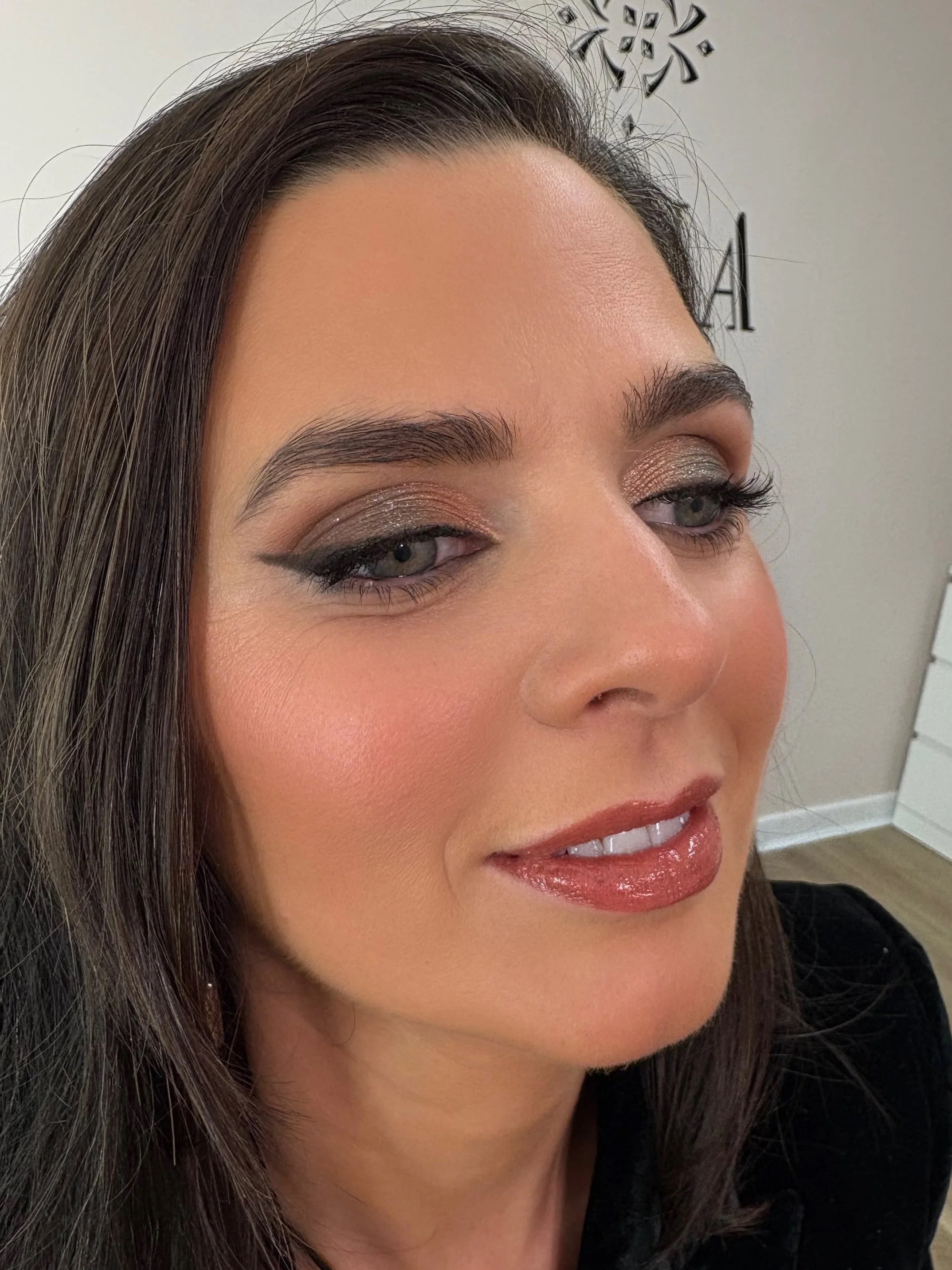 Close-up of a woman's face with styled hair, makeup including eye shadow, eyeliner, and lipstick, smiling, with part of a wall with a decorative element and some text in the background.