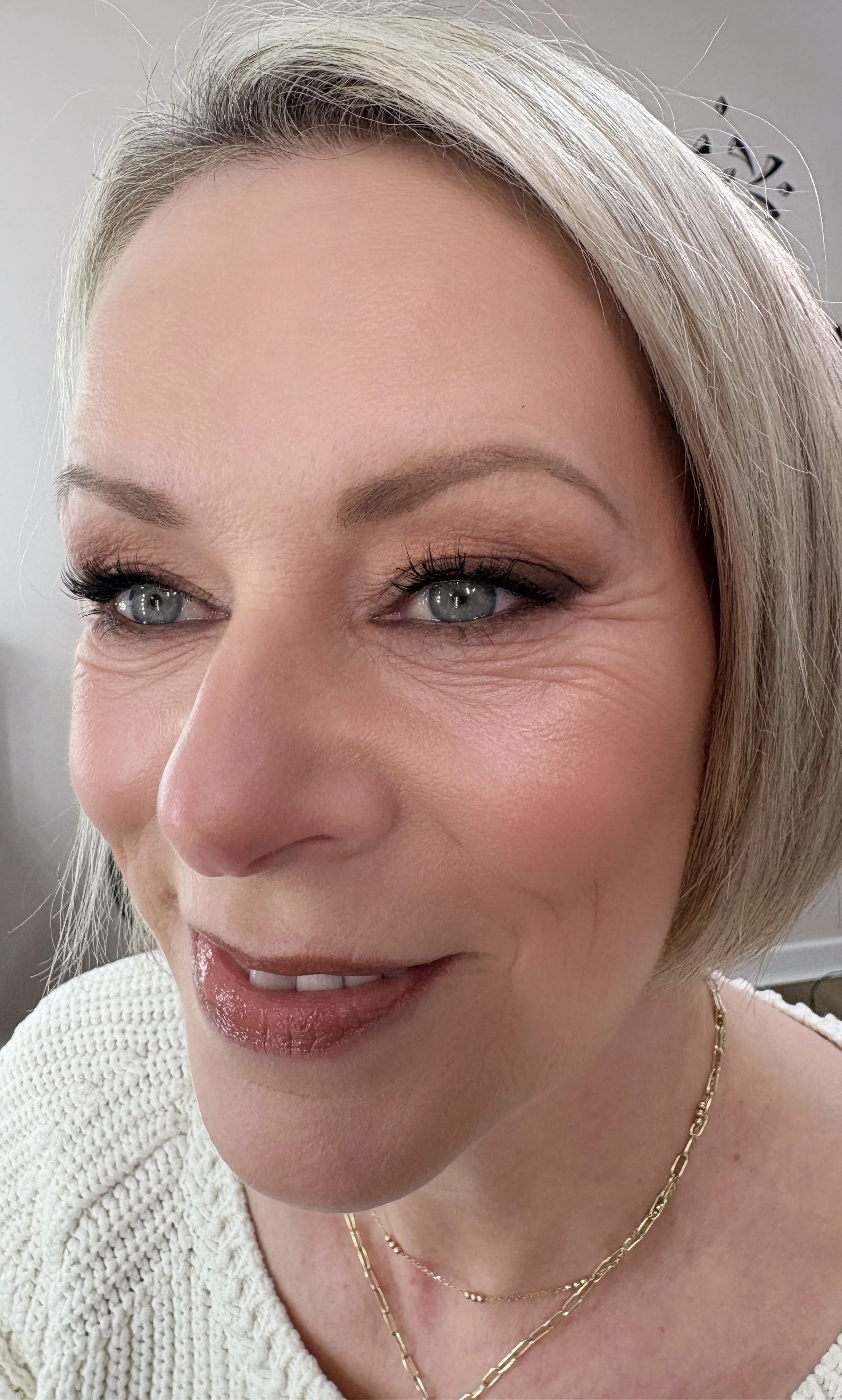 Close-up of a woman's face, showing her eyes, nose, lips, and part of her blonde hair, with jewelry around her neck, wearing makeup, and a white knitted sweater.