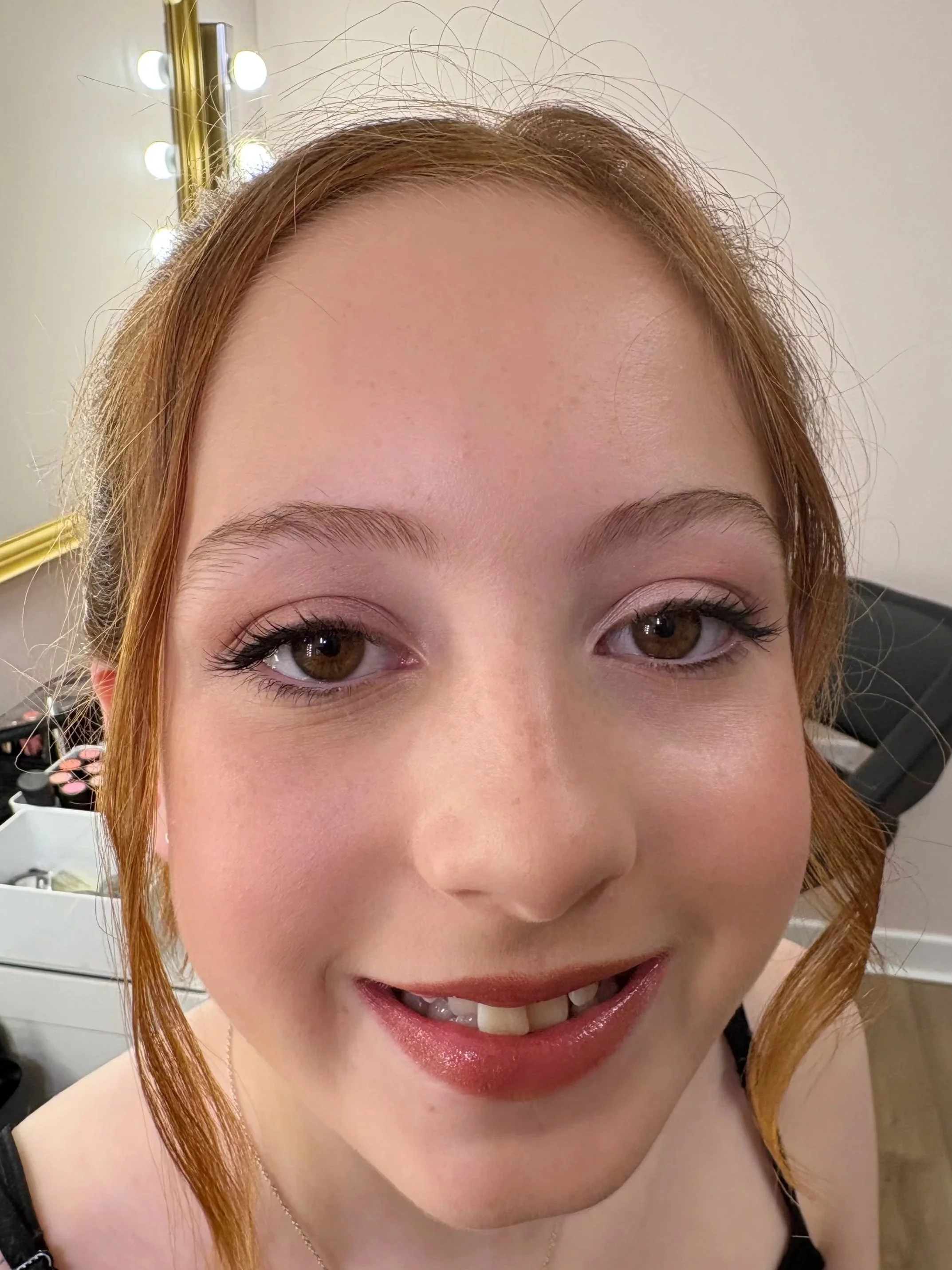 A close-up photo of a woman with red hair, smiling, showing her teeth, with makeup, in a room with a mirror and makeup items in the background.