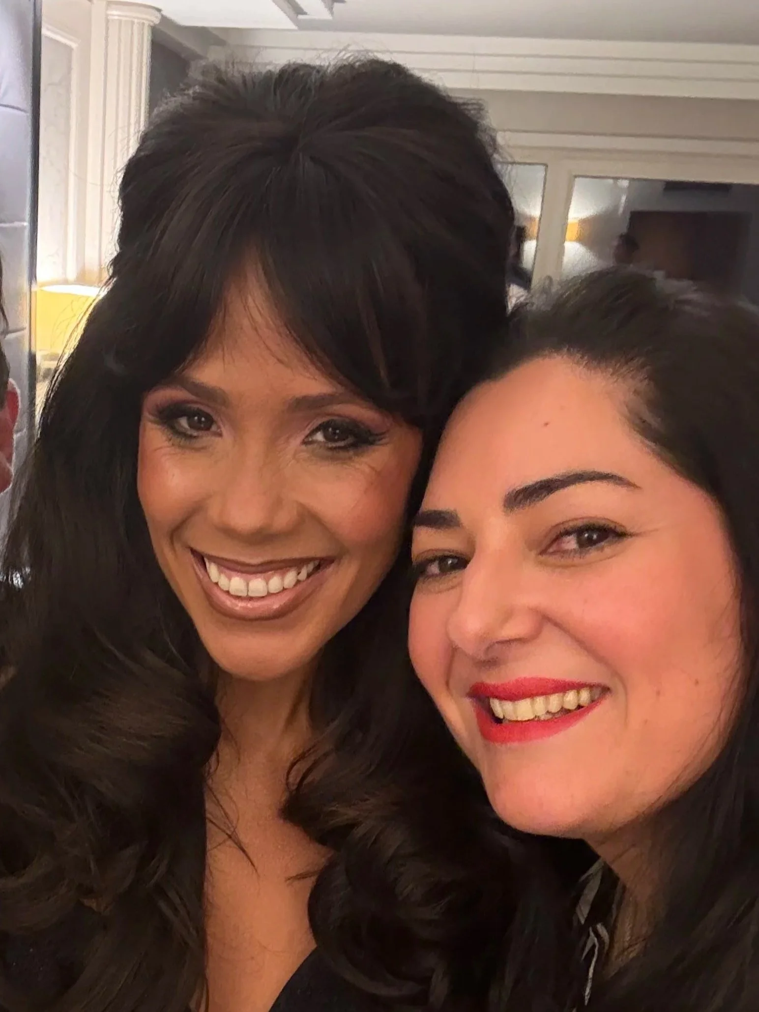 Close-up of two women smiling with long dark hair, one with dark lipstick, in an indoor setting.