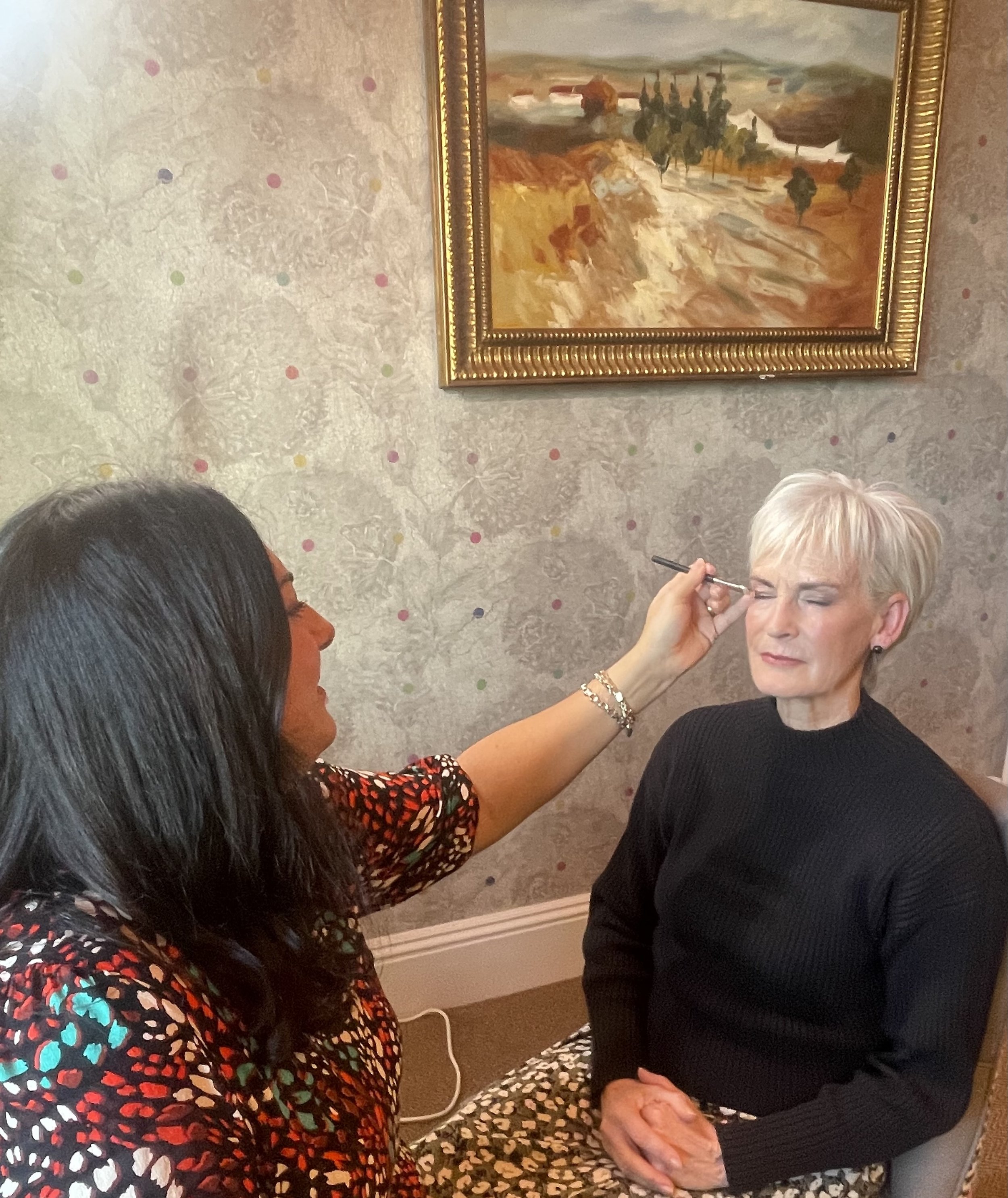 A woman with painted eyebrows and closed eyes is having her makeup done by another person in a room with textured wallpaper, a framed painting, and a floral chair.