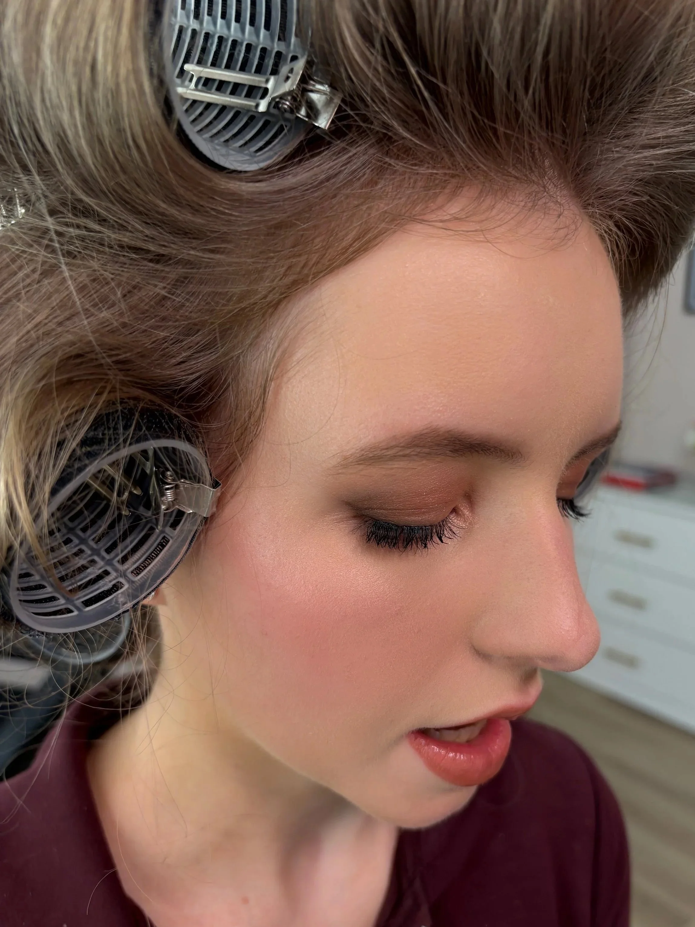Close-up of a woman's face with blonde hair, wearing a hair roller or diffuser on her head, with her eyes closed and smiling.