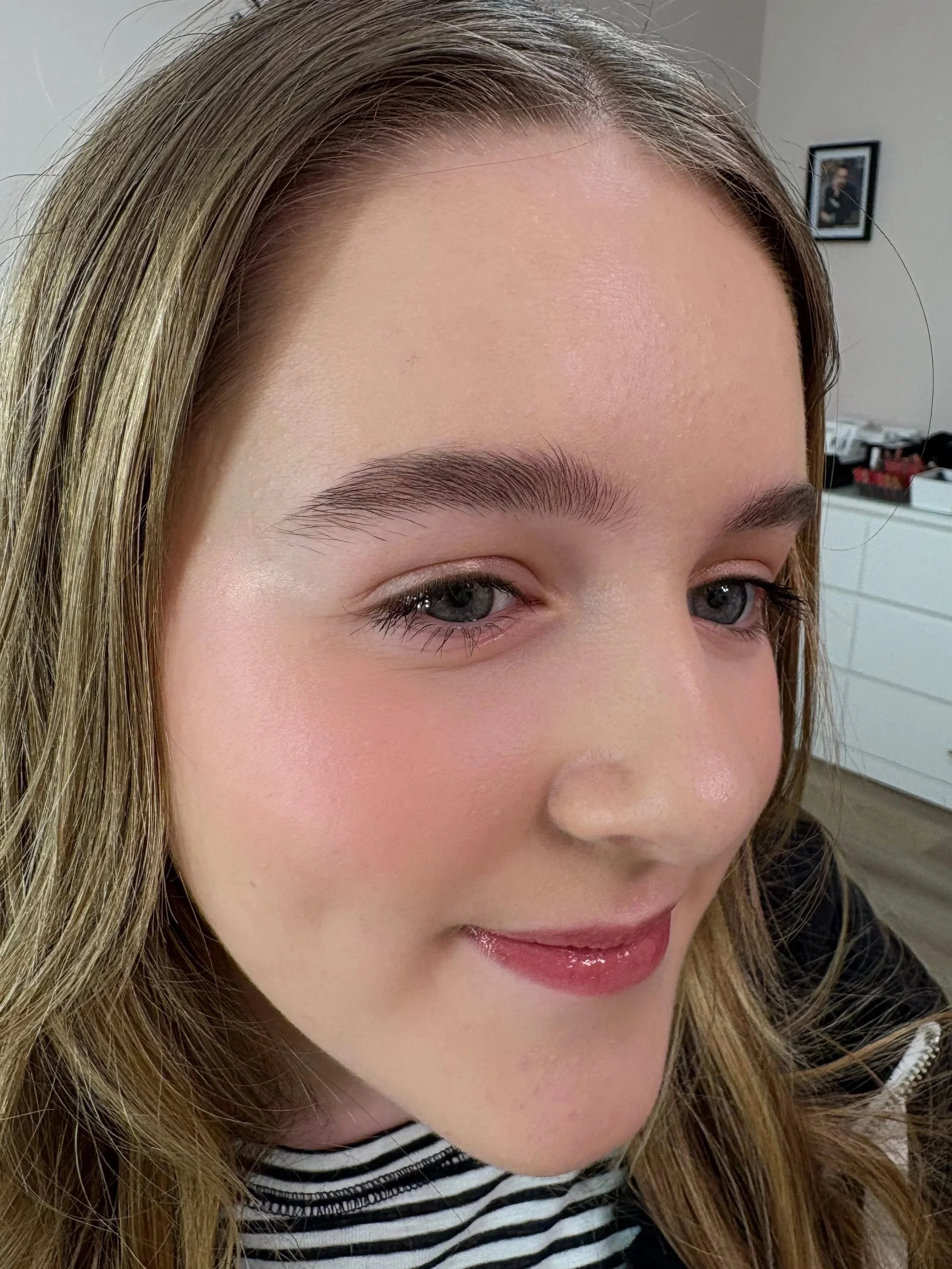 Close-up of a woman with blonde hair, blue eyes, and glossy pink lips, smiling softly. She is wearing a black and white striped shirt, and a framed photo is visible in the background.