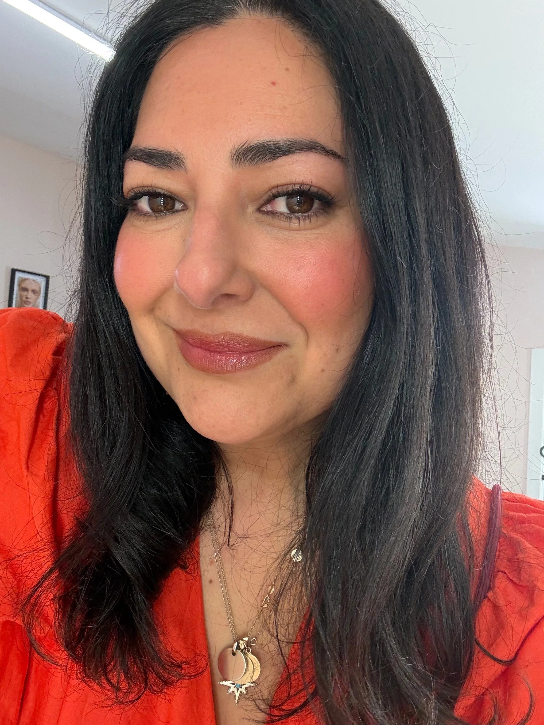 A woman with long black hair and brown eyes wearing a red top and layered necklaces, smiling and looking at the camera. A framed picture of a woman is visible in the background.