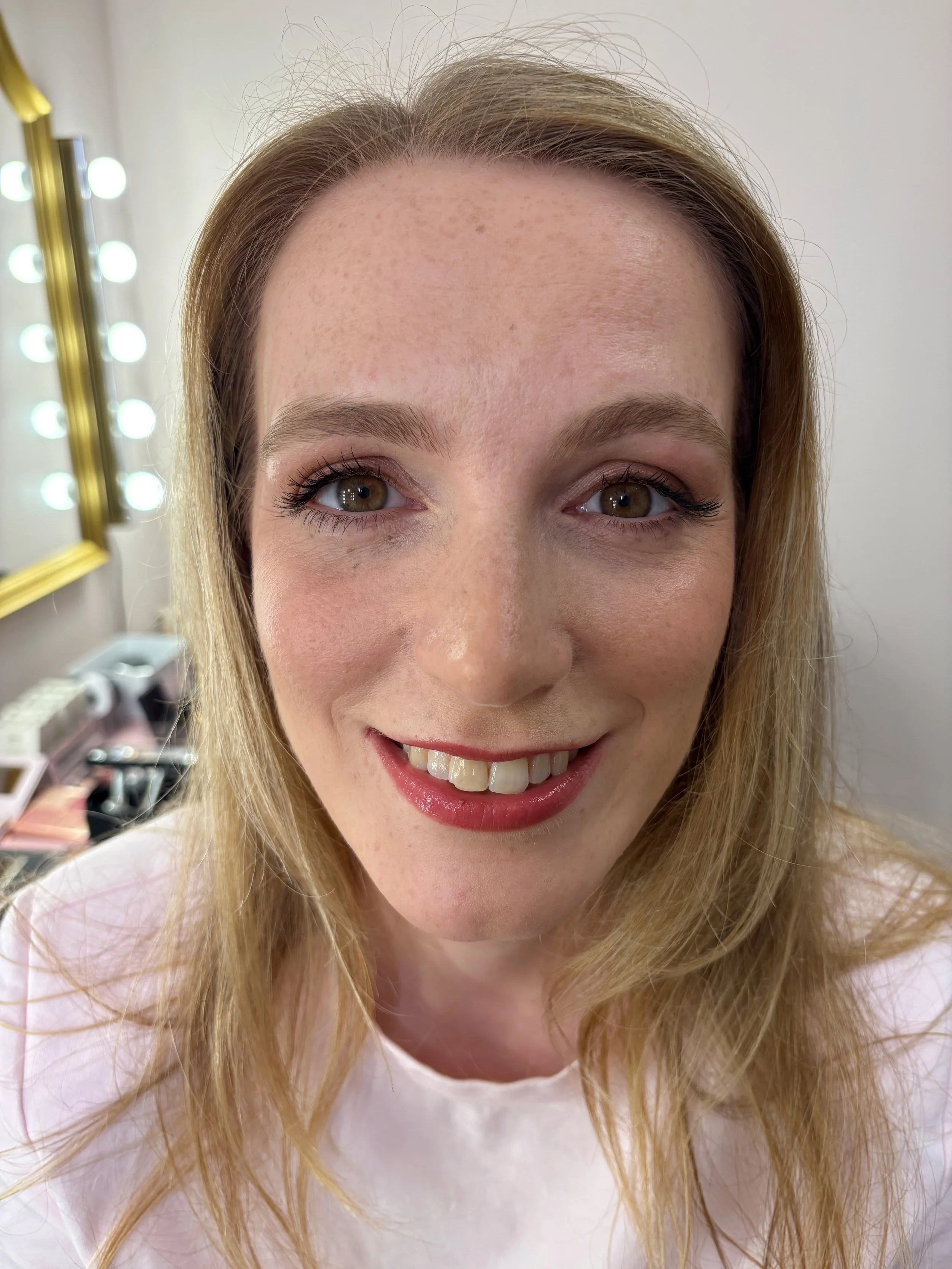 A woman with light skin, freckles, and wavy blonde hair smiling, with makeup including eyeshadow, eyeliner, mascara, and lipstick. The background shows a mirror with lights and makeup products.