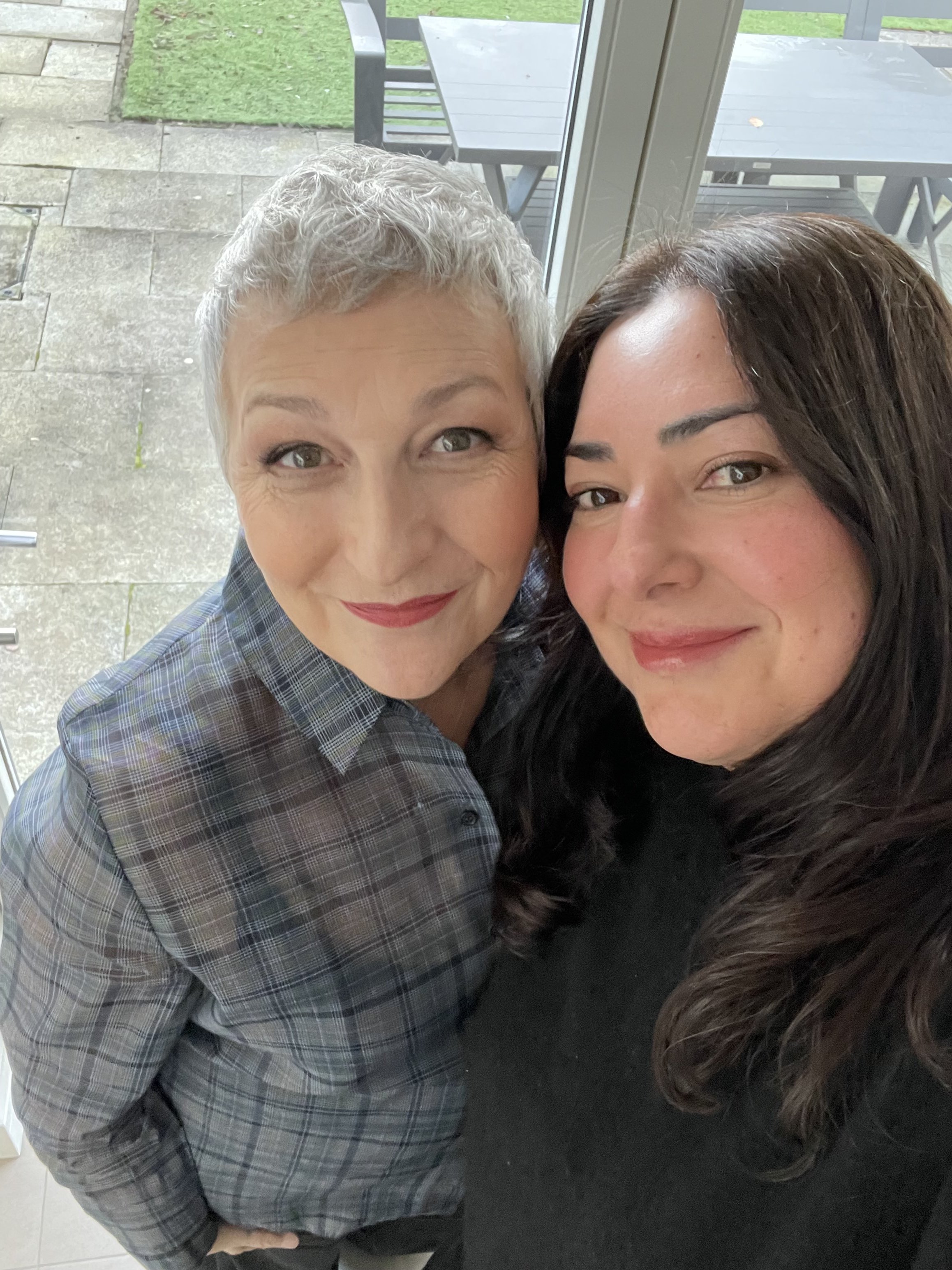Close-up of two smiling women taking a selfie together, one with short curly gray hair and the other with long dark hair, inside a building near a glass door leading to an outdoor patio with tables and chairs.