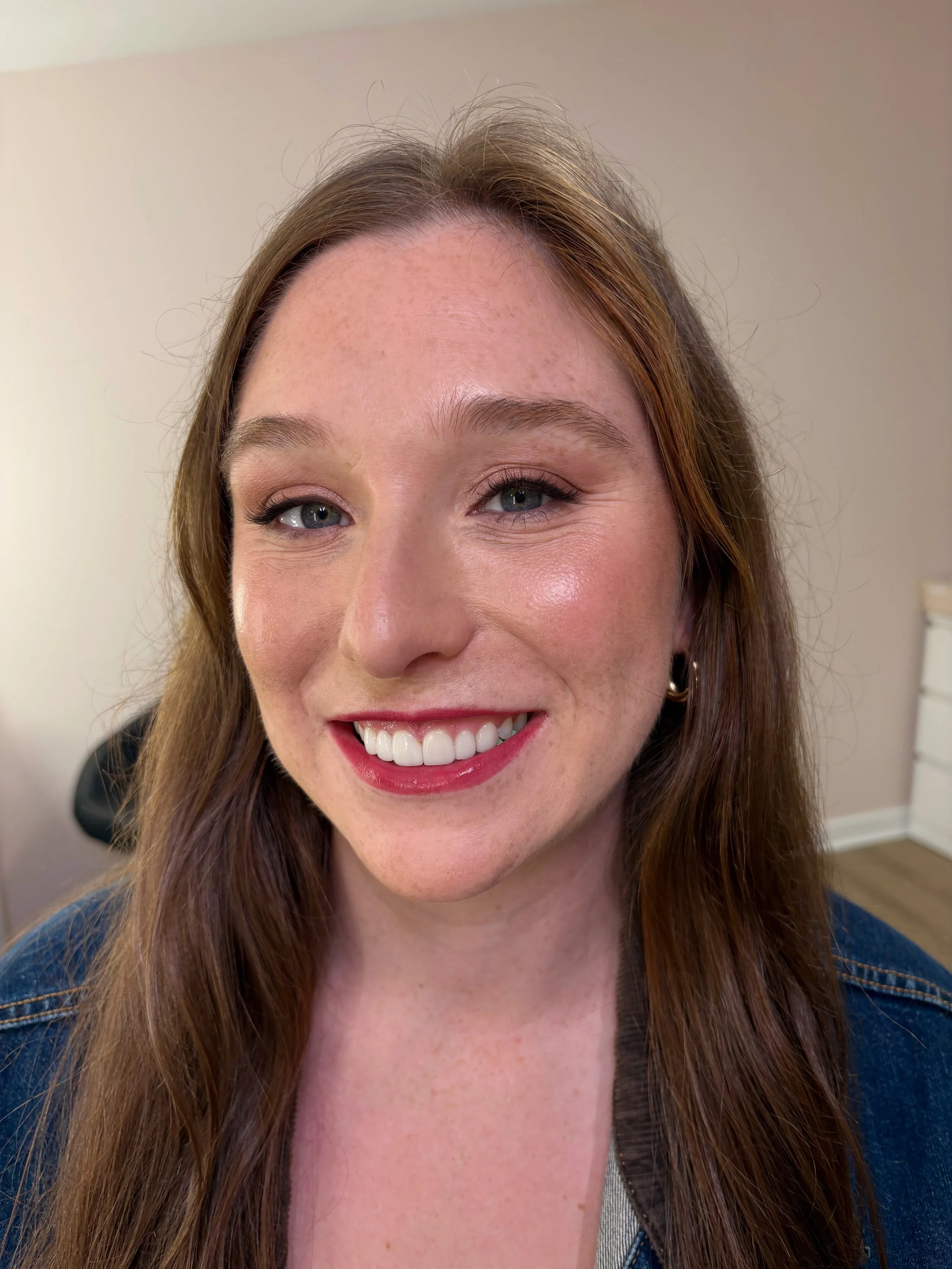 Close-up of a woman with light skin, long brown hair, blue eyes, smiling, wearing hoop earrings and a denim jacket, indoor setting with neutral background.