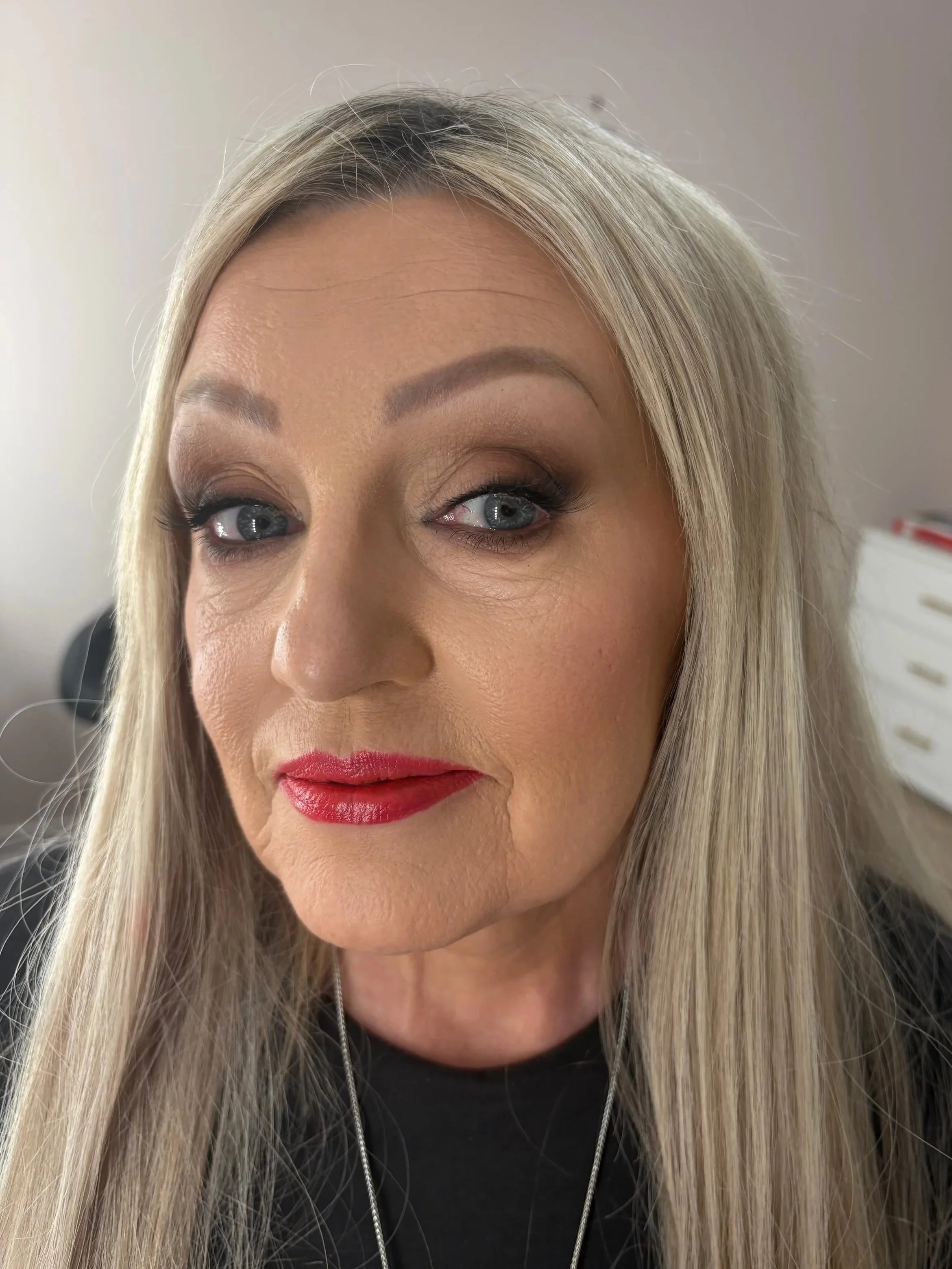 Close-up of a woman with long blonde hair, blue eyes, wearing makeup including dark eyeshadow, eyeliner, mascara, and red lipstick, with a neutral background.