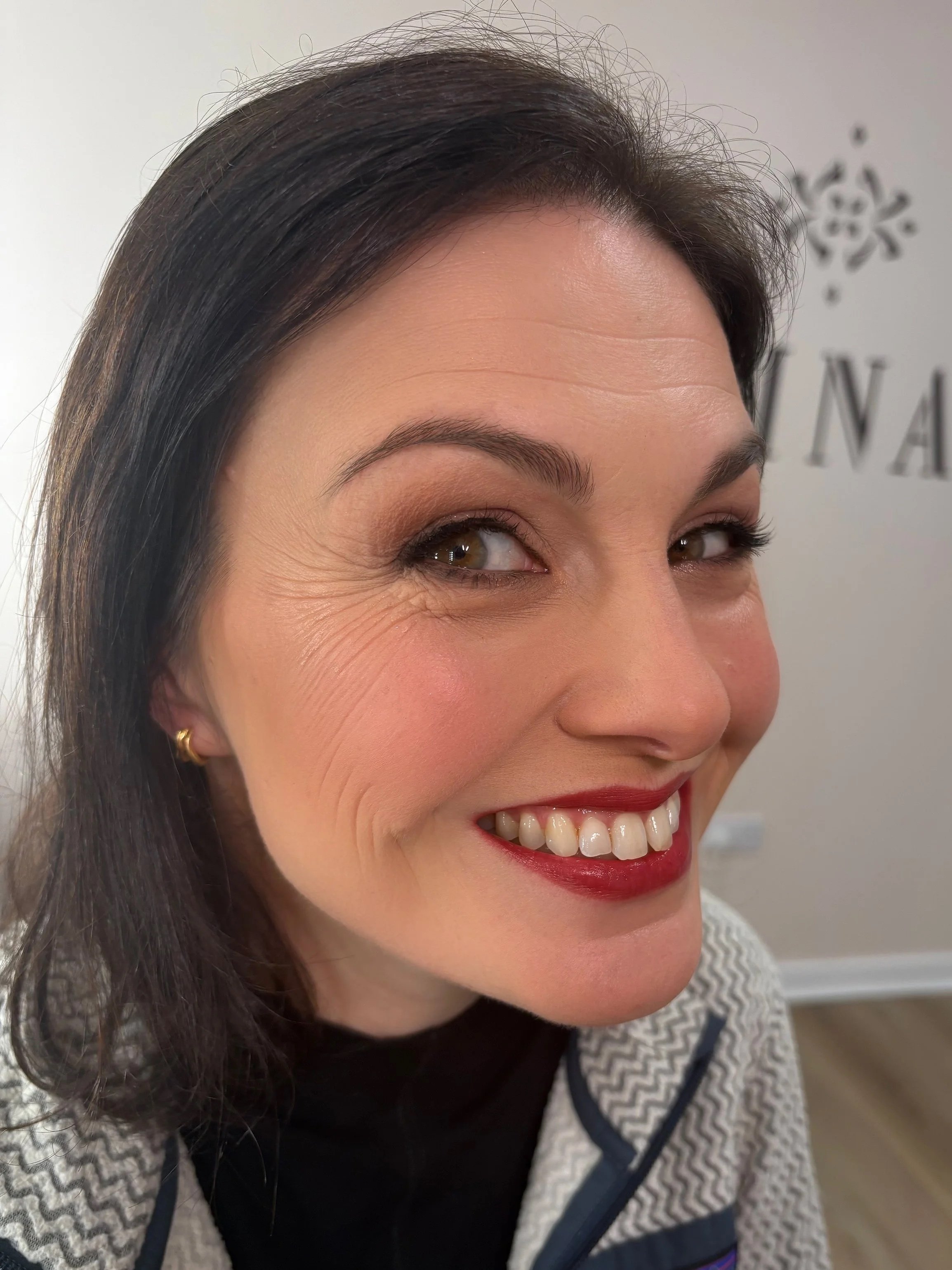 Close-up of a smiling woman with dark hair, wearing a patterned sweater, small gold earrings, and red lipstick, with a wall and partial text in the background.