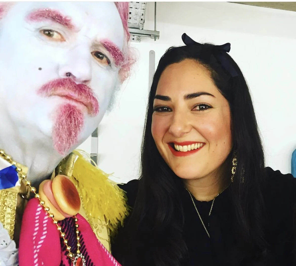 A woman with long black hair, red lipstick, and a black bow headband smiling next to a man with white face paint, pink makeup, and colorful clothing, both taking a selfie together.