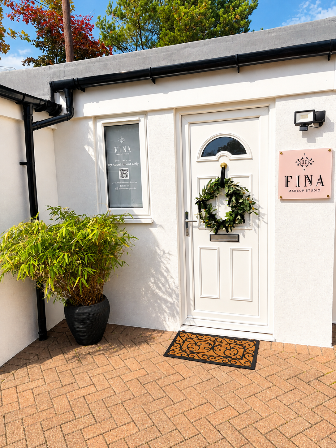 White door with a wreath, a window with text, a pink sign reading 'FINA Makeup Studio,' a black potted plant, and a decorative doormat outside a building on a brick patio, under a blue sky with green trees.