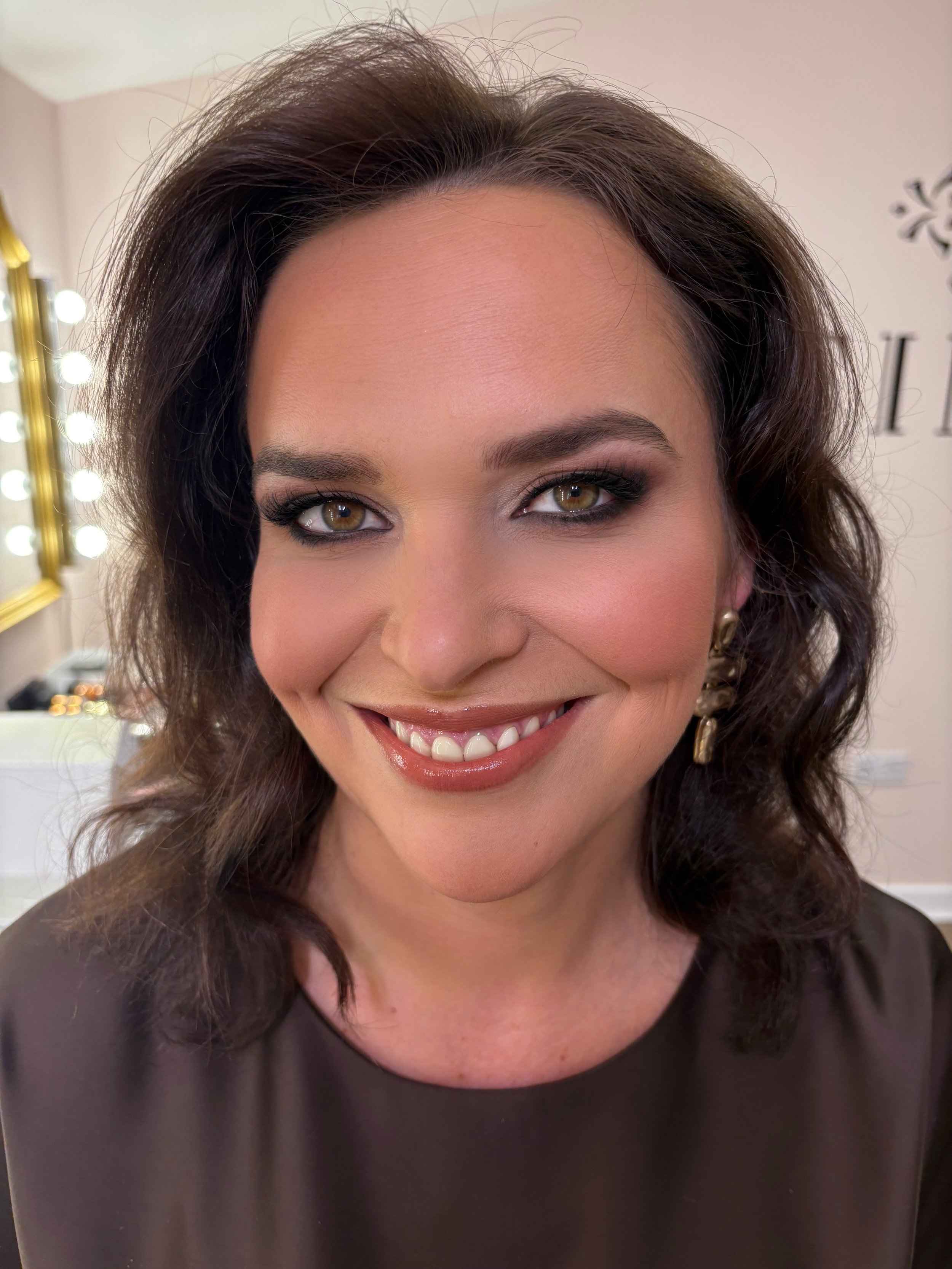 Close-up of a smiling woman with brown hair and hazel eyes, wearing makeup and earrings, in a well-lit room.