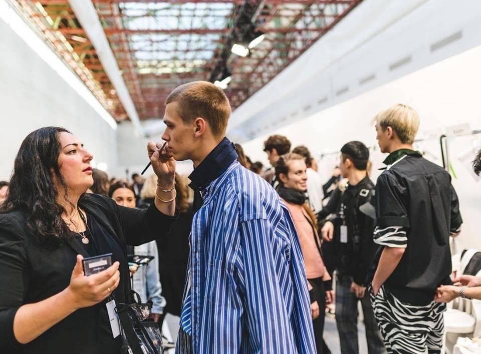 A makeup artist applying makeup to a young man at a busy event.
