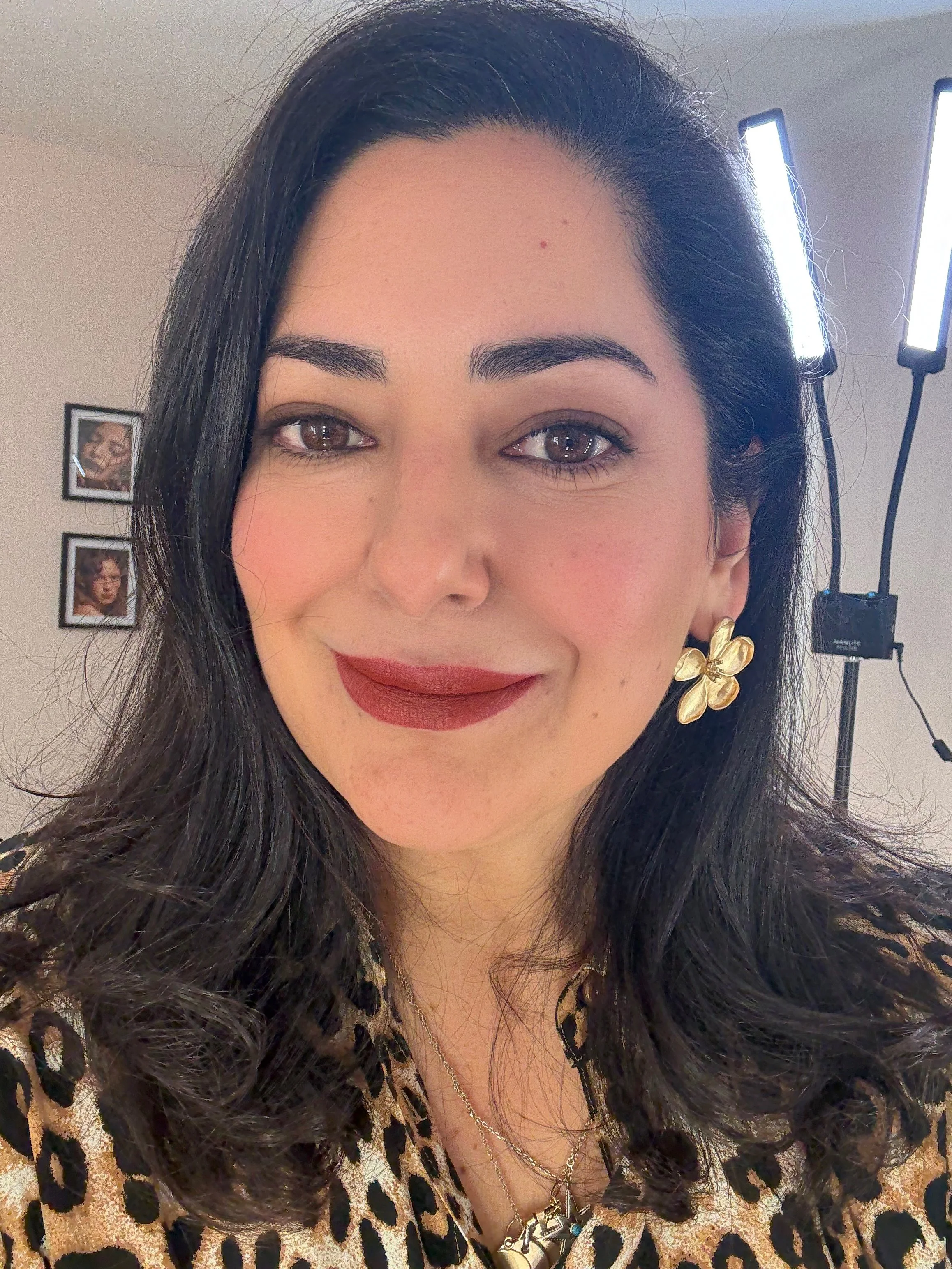 A woman with dark hair, wearing a leopard print top, gold flower earrings, and layered necklaces, smiling in front of a backdrop with bright lights and framed artwork on the wall.