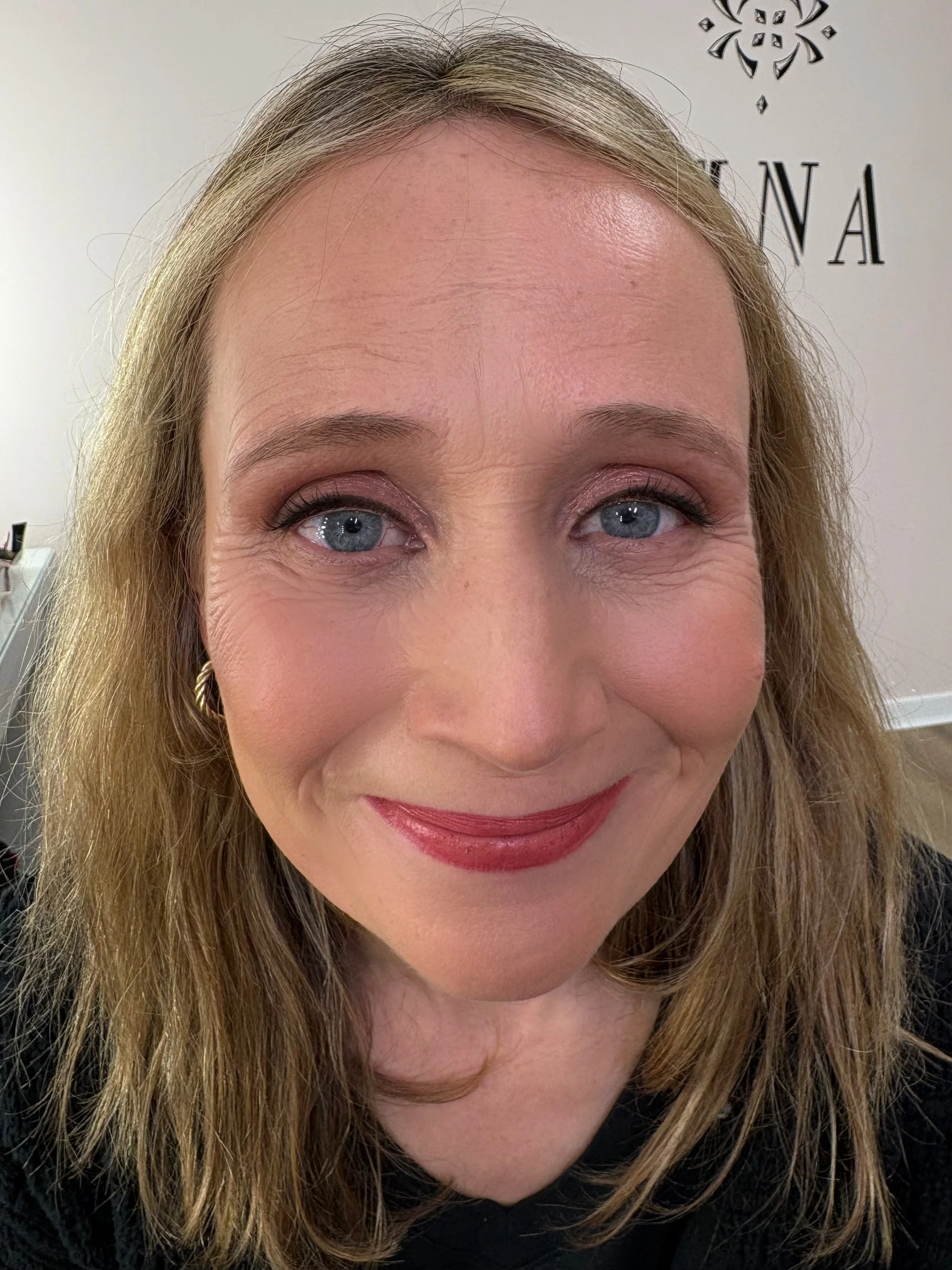 A woman with blonde hair and blue eyes, smiling with lipstick, looks at the camera. Background features part of a room with a sign or logo on the wall.