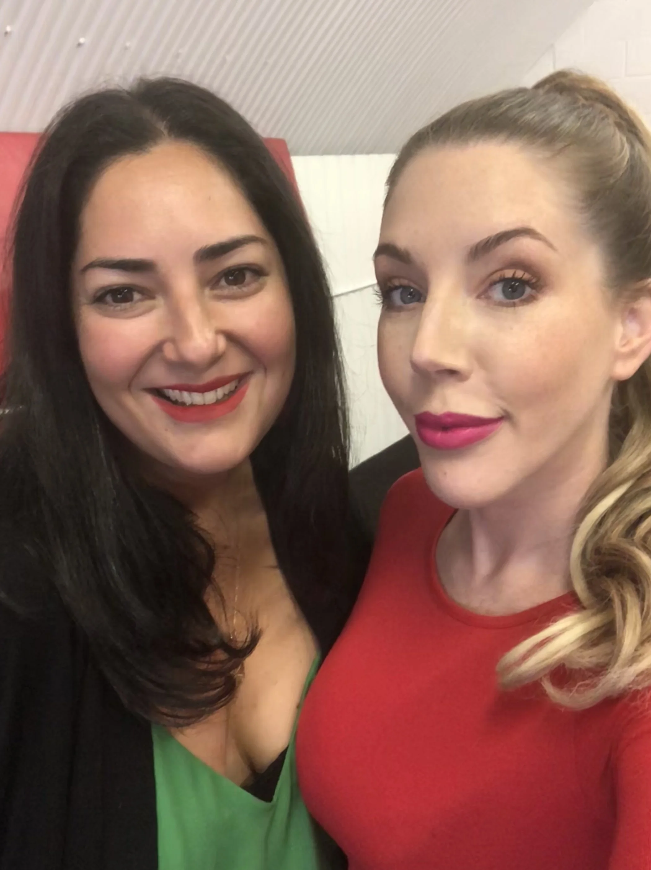 Two women smiling, one with black hair wearing a green top, the other with blonde hair wearing a red top, with a curved ceiling in the background.