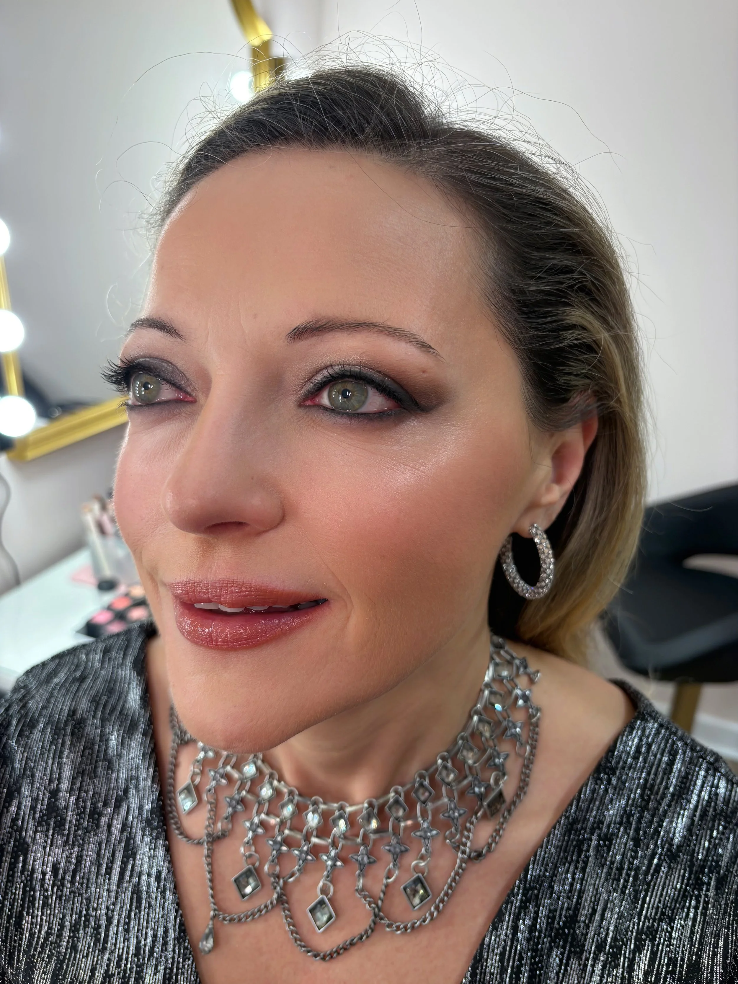 Close-up of a woman's face showing her eyes, makeup, earrings, and layered silver necklaces, with a mirror and makeup products in the background.
