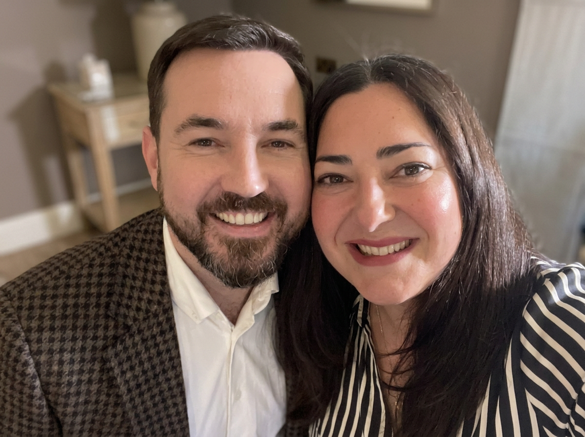 A smiling man and woman taking a close-up selfie indoors, with a blurred background showing a small table and decorative items.