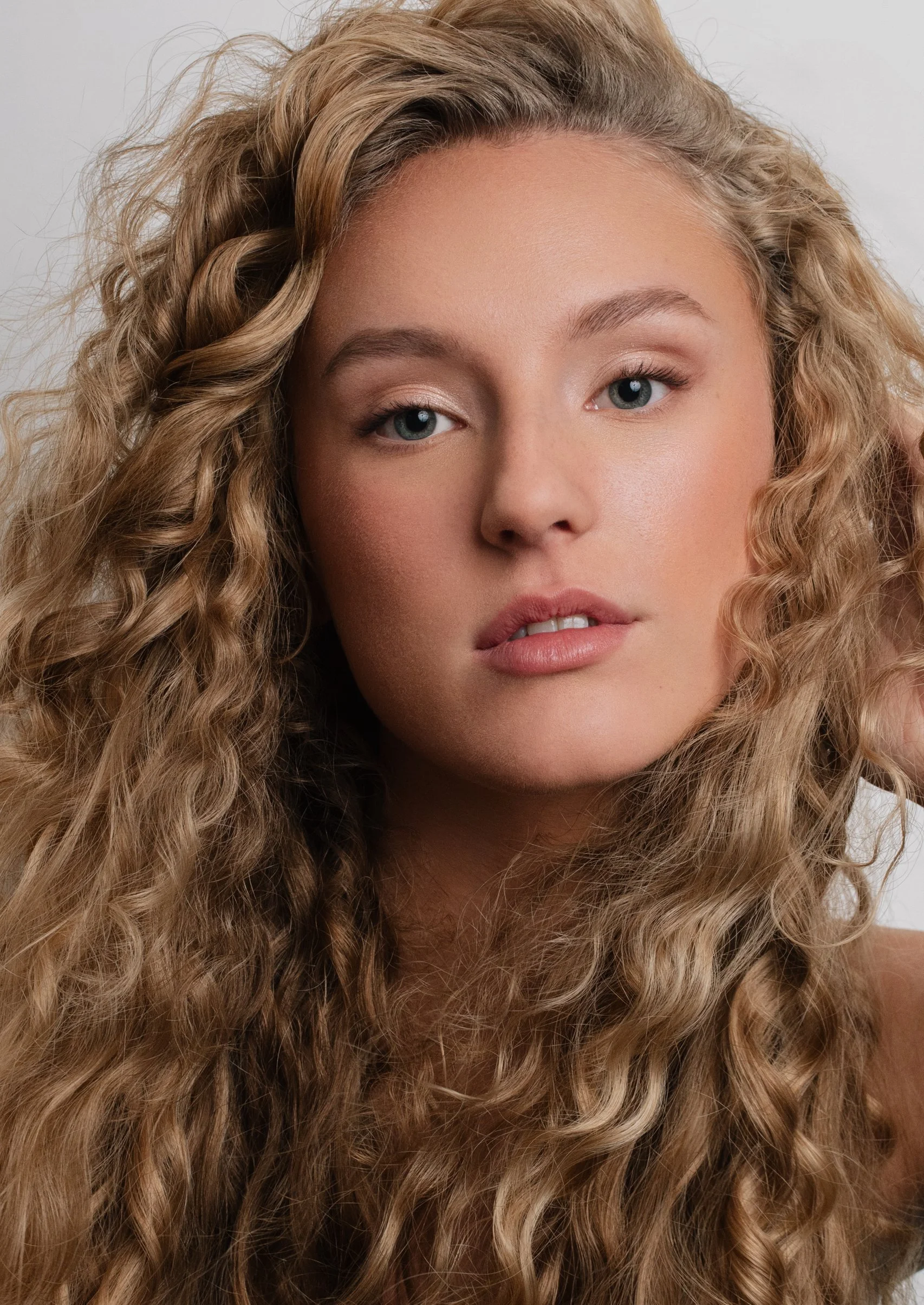 Close-up of a woman's face with curly blonde hair, blue eyes, and natural makeup.