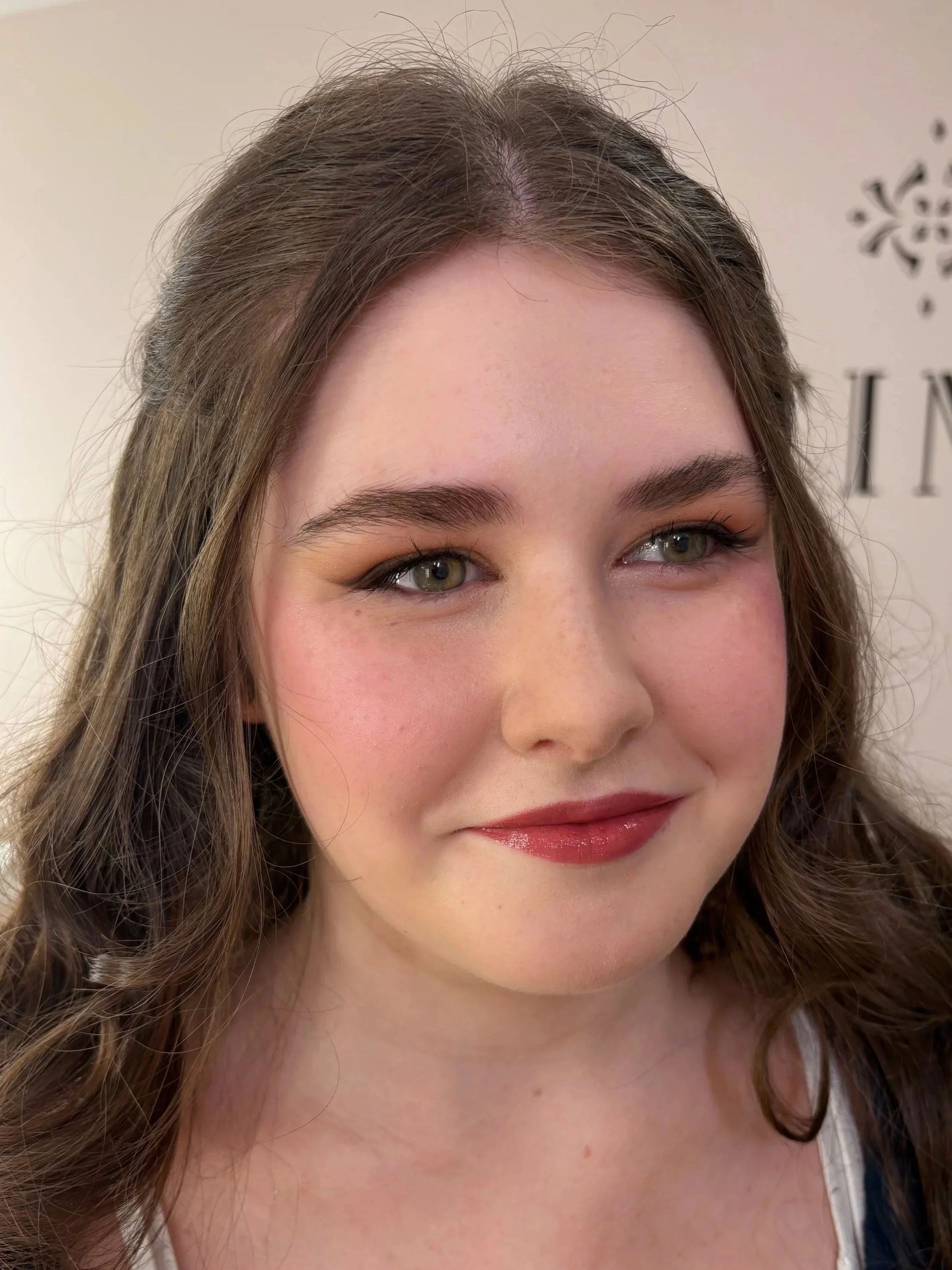 Close-up of a smiling young woman with curly brown hair, blue eyes, and natural makeup, wearing a white top.