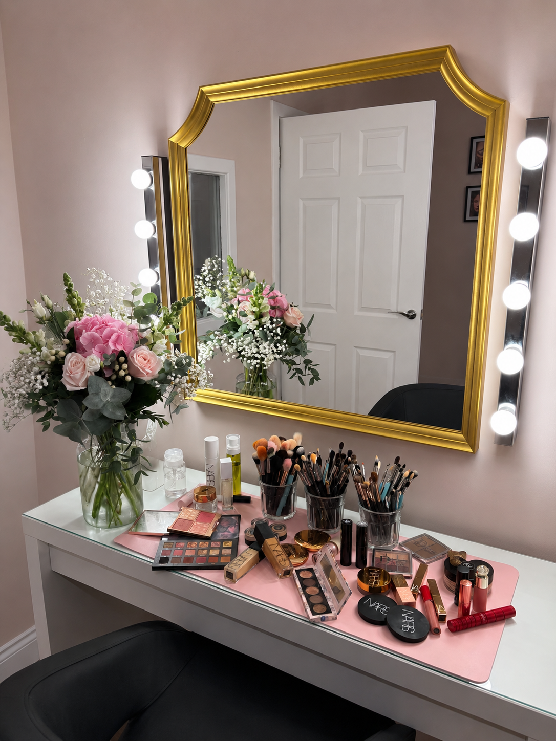 A makeup vanity table with a large mirror framed in gold, illuminated by bulbs on either side. The table has a pink mat with various makeup products including brushes, lipsticks, palettes, and skincare bottles. There is a large bouquet of pink and white flowers in a glass vase to the left of the table.