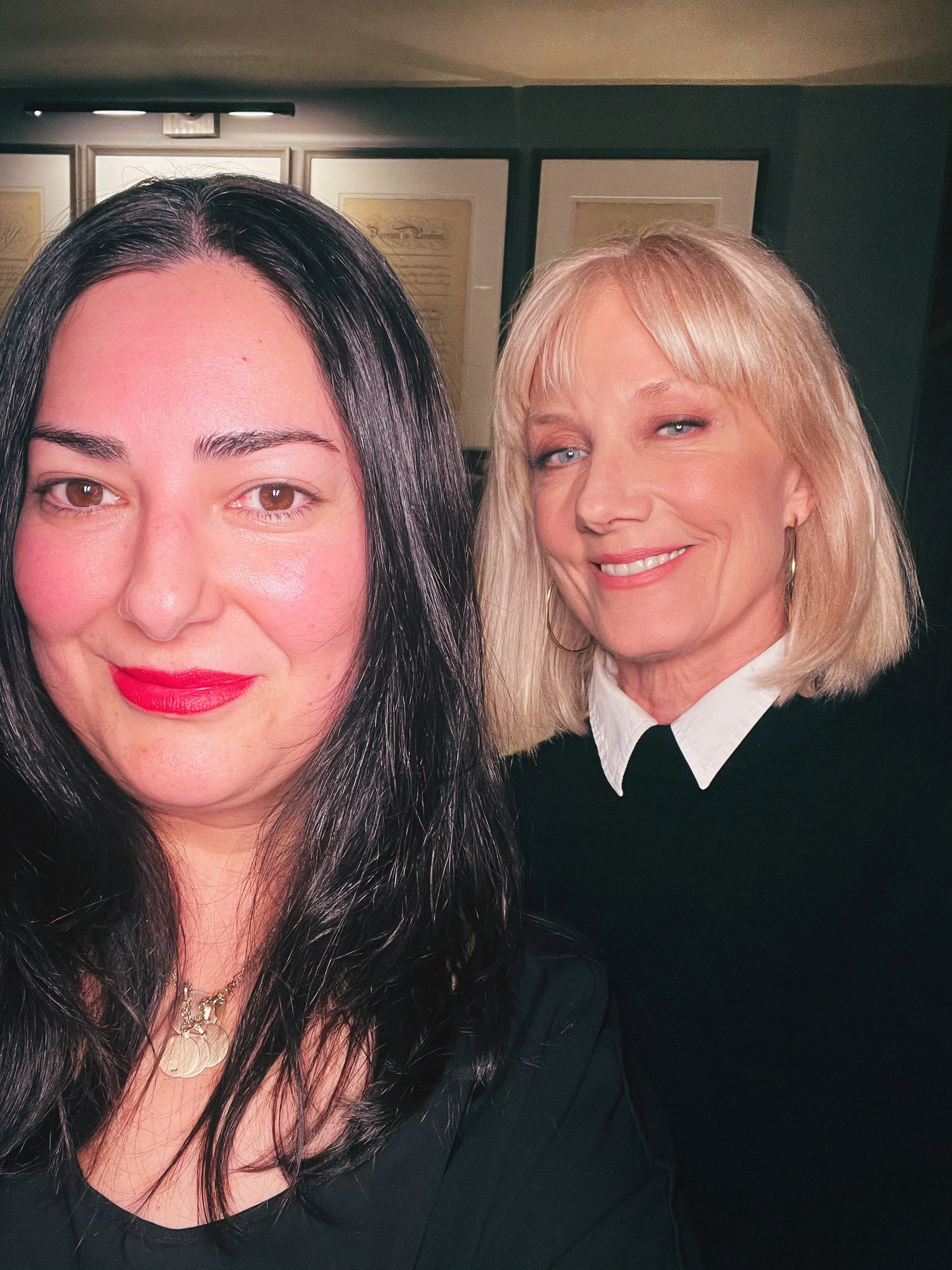 Close-up of two women smiling, with the woman on the left having long black hair and the woman on the right having shoulder-length blonde hair, in front of framed artwork on a dark wall.