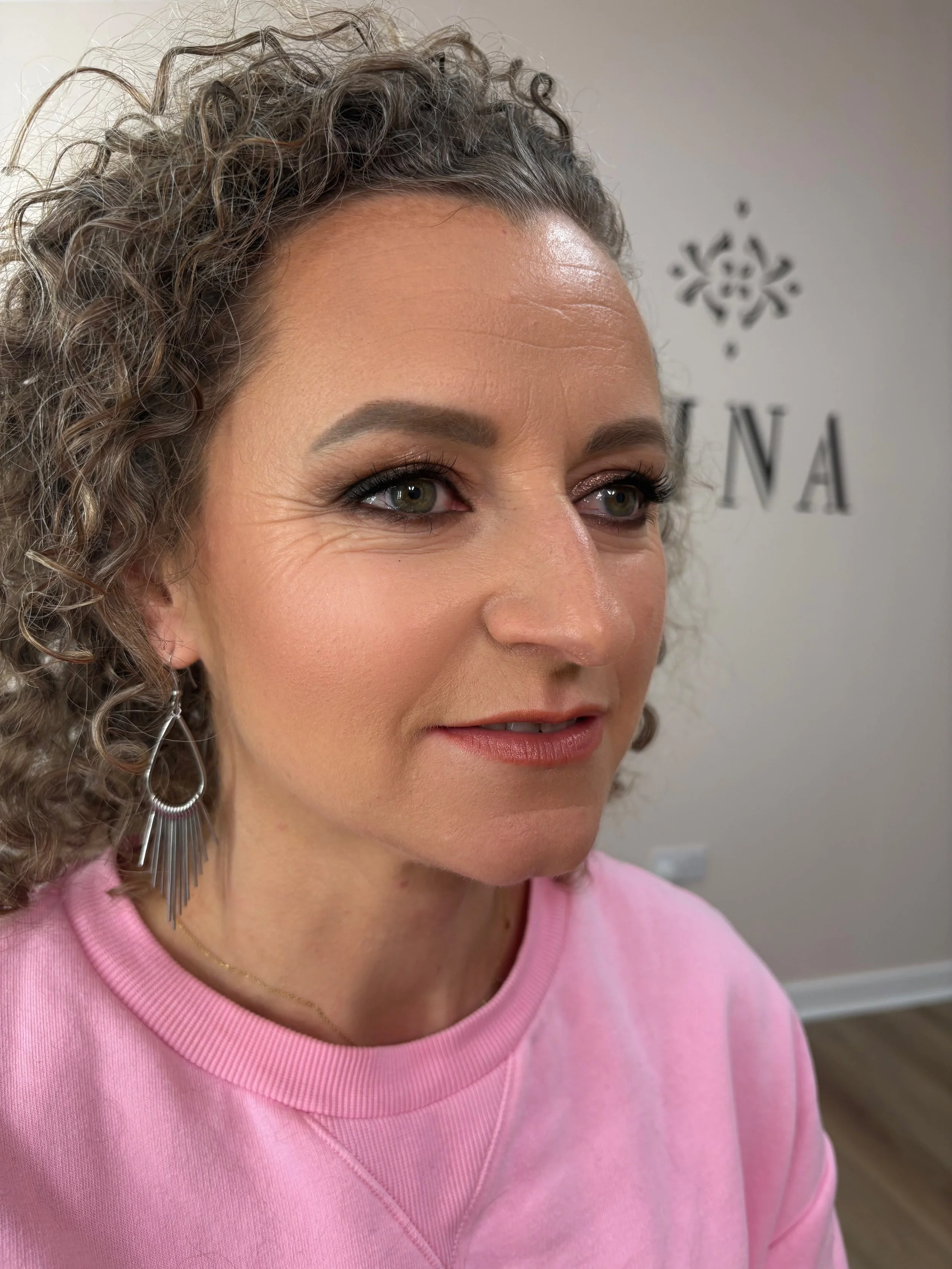 Close-up of a woman with curly gray hair, wearing a pink top and silver earrings, smiling softly in front of a wall with a decorative pattern and the letters 'NA'.