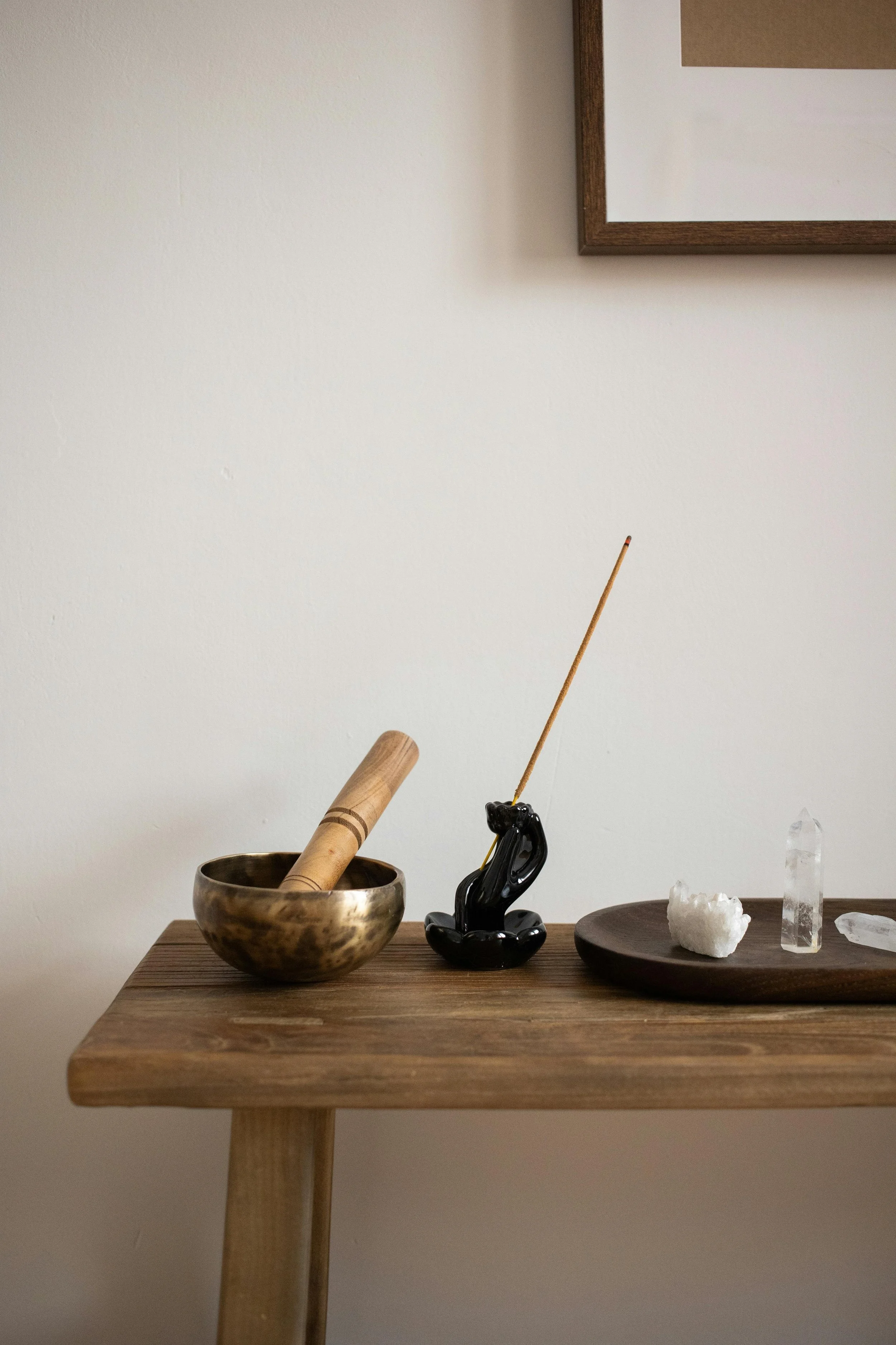 A wooden table with a brass singing bowl containing a wooden mallet, a black abstract sculpture, a white mineral specimen, and a tall clear crystal point. A framed picture hangs on the wall above.