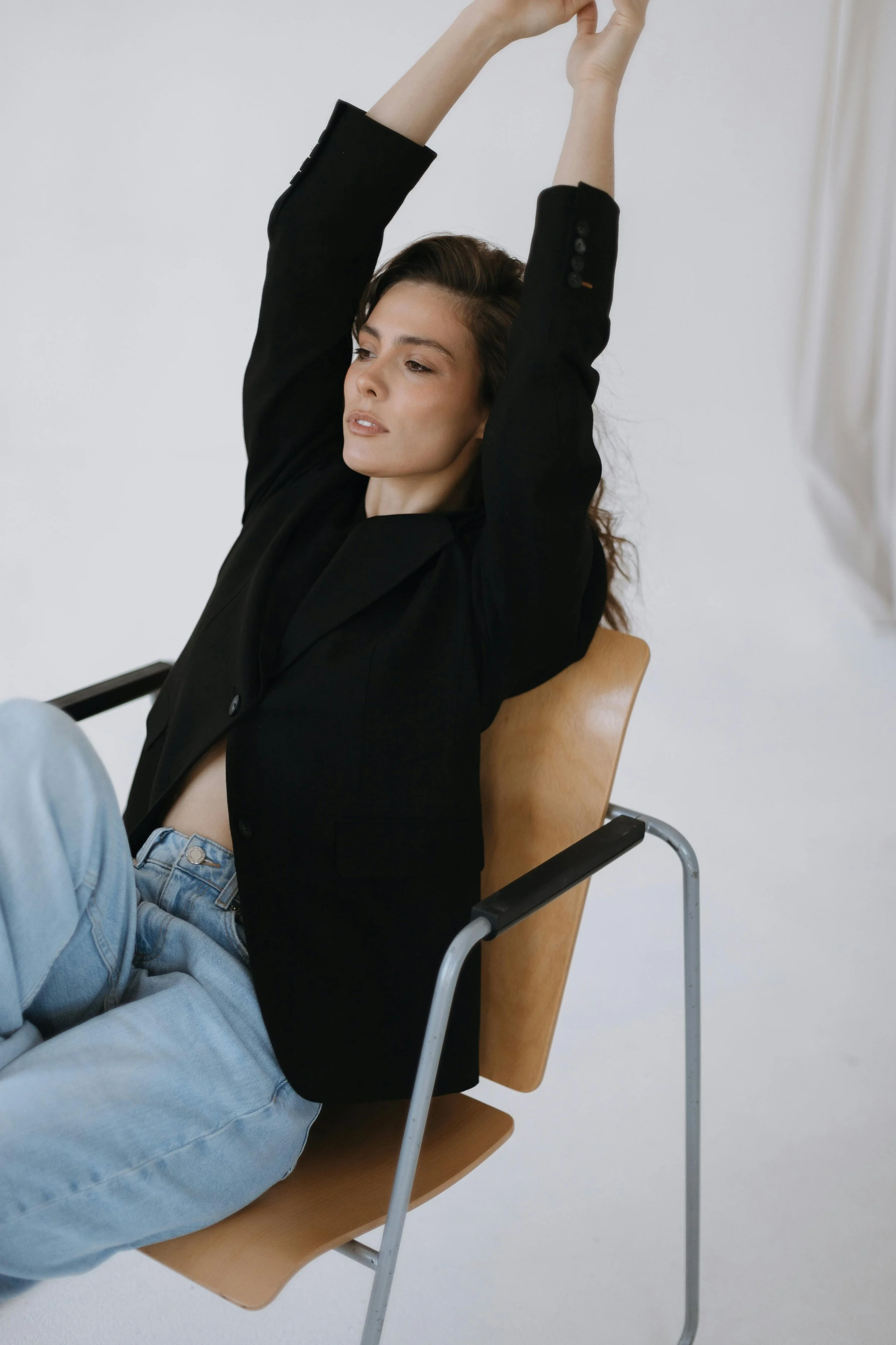 A woman with long hair sitting on a wooden chair with her arms raised above her head, wearing a black blazer and light blue jeans, against a plain white wall.