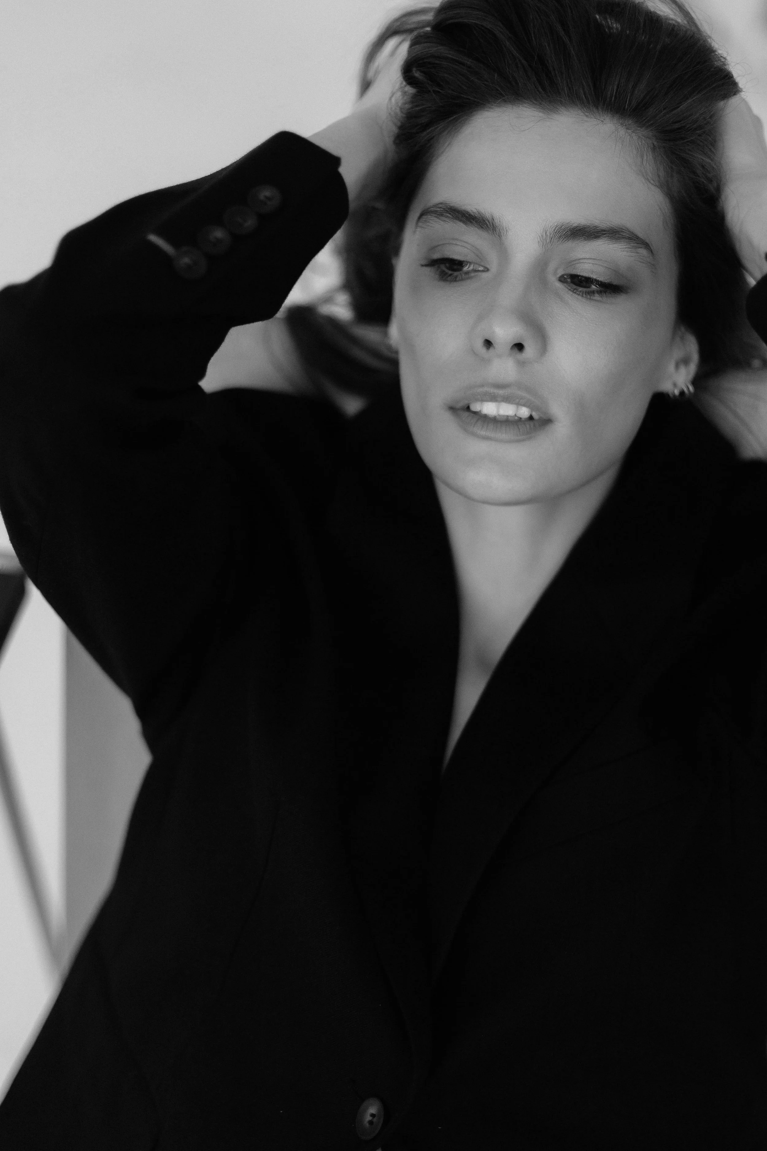 A woman with dark hair, parted to the side, laying her head back with her hands on her head. She is wearing a black blazer with button details on the sleeve.