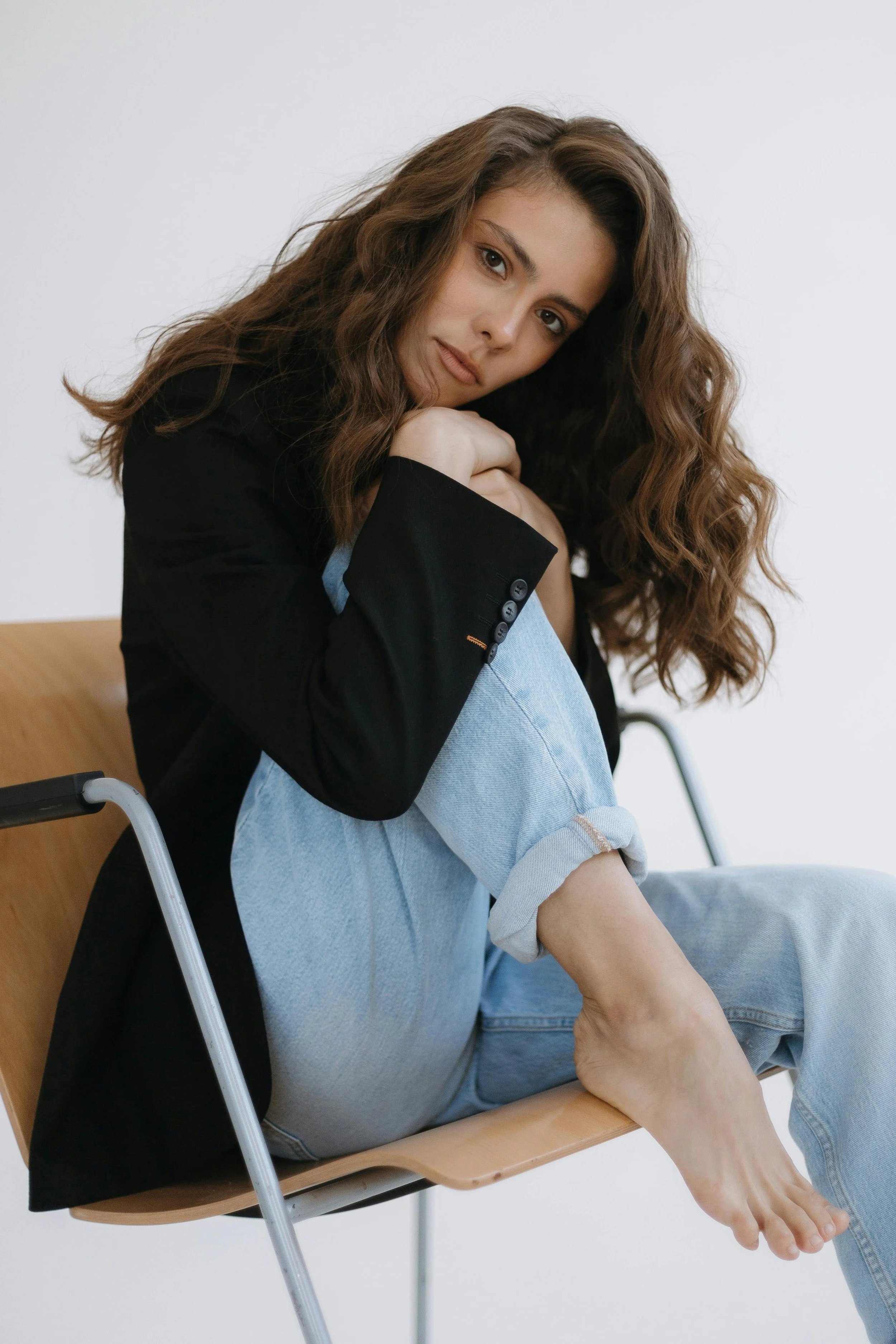 A woman sitting in a chair with a wooden desk attached, resting her cheek on her hands, looking at the camera with a neutral expression, wearing a black blazer and light blue jeans, with plain white background.