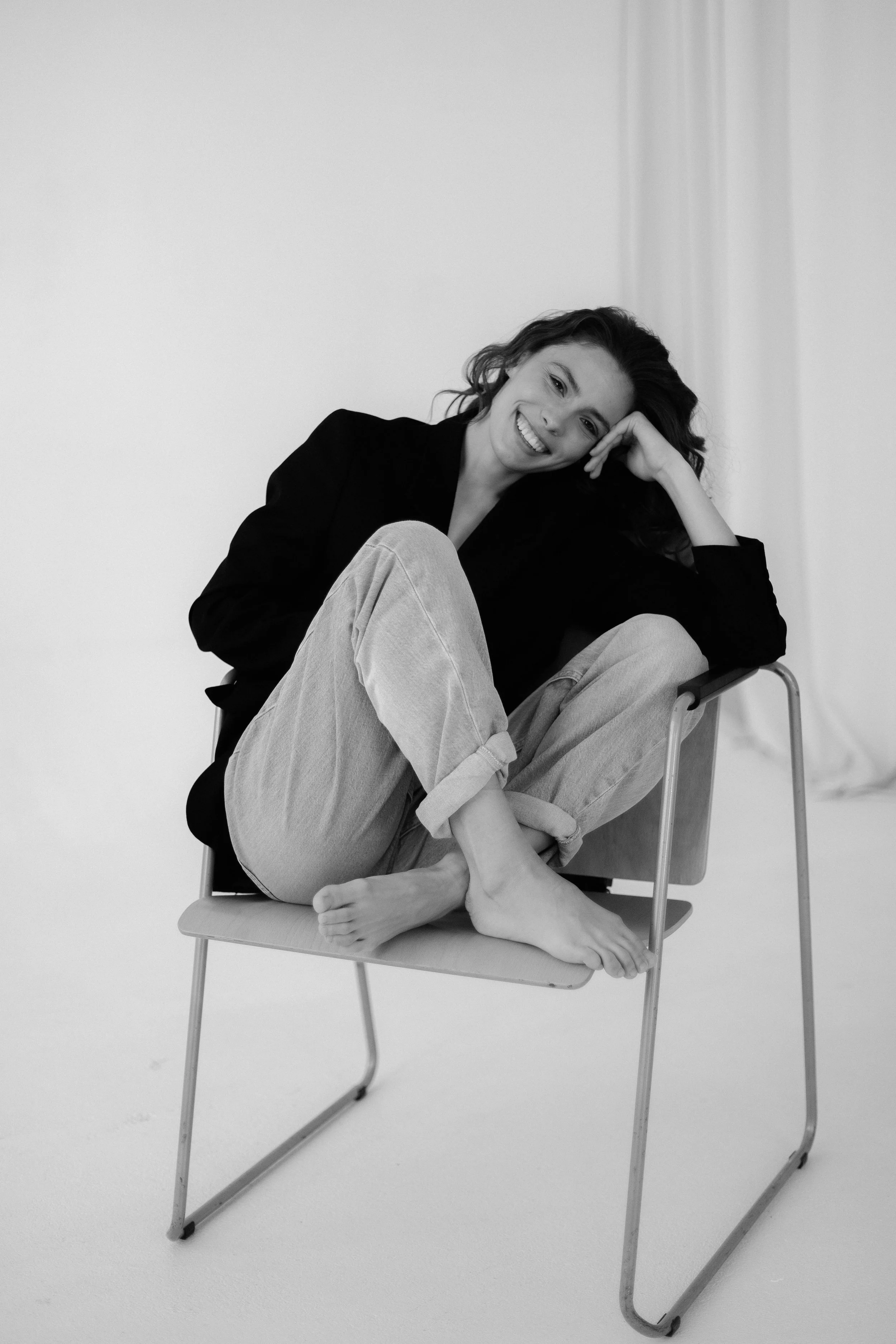 Black and white photograph of a young woman sitting barefoot on a chair, smiling, with one leg crossed over the other. She is wearing a dark blazer and light pants, with her head resting on her hand.