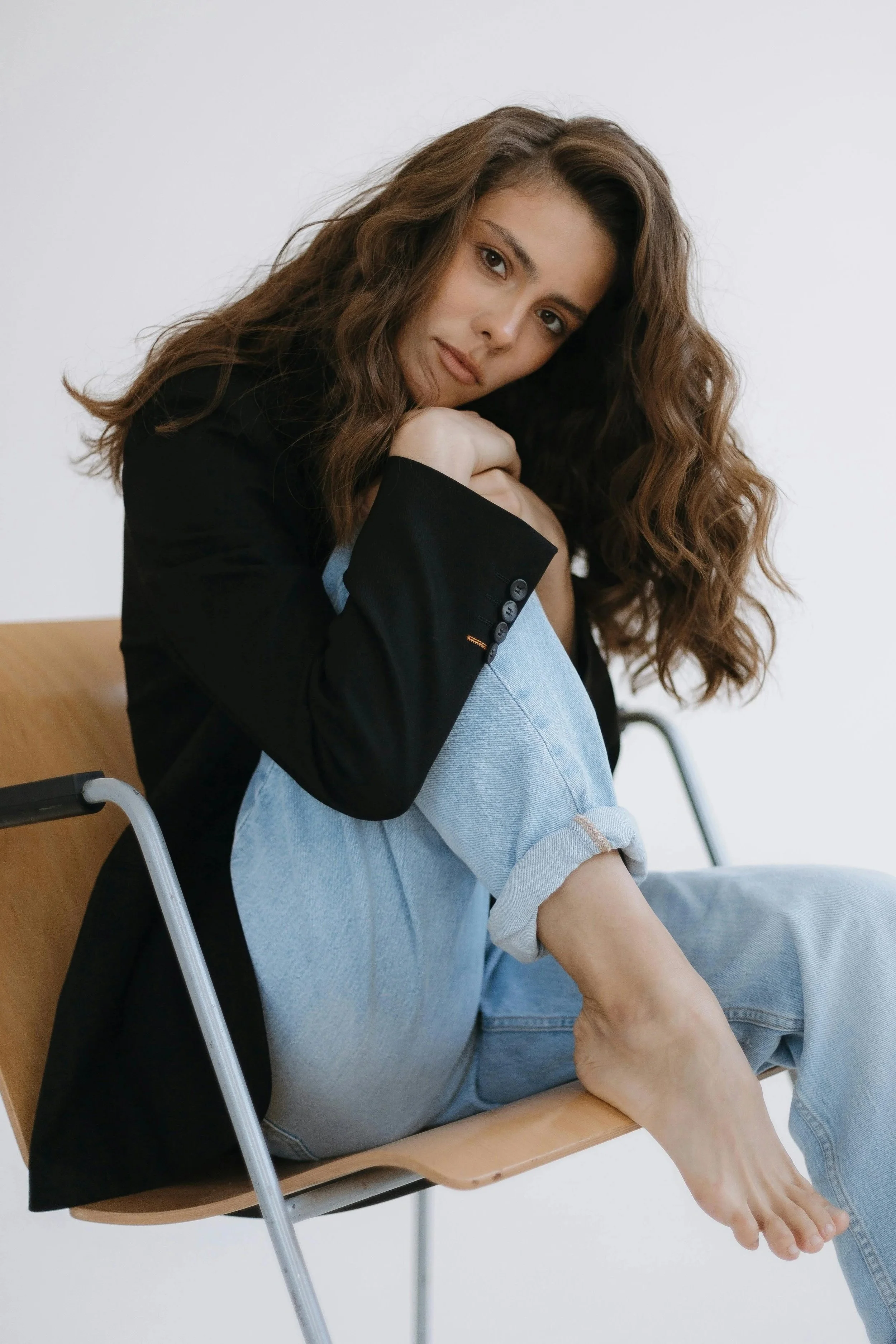 A young woman with curly brown hair sitting at a desk, resting her head on her folded arms, wearing a black jacket and light blue jeans, with a plain white background.