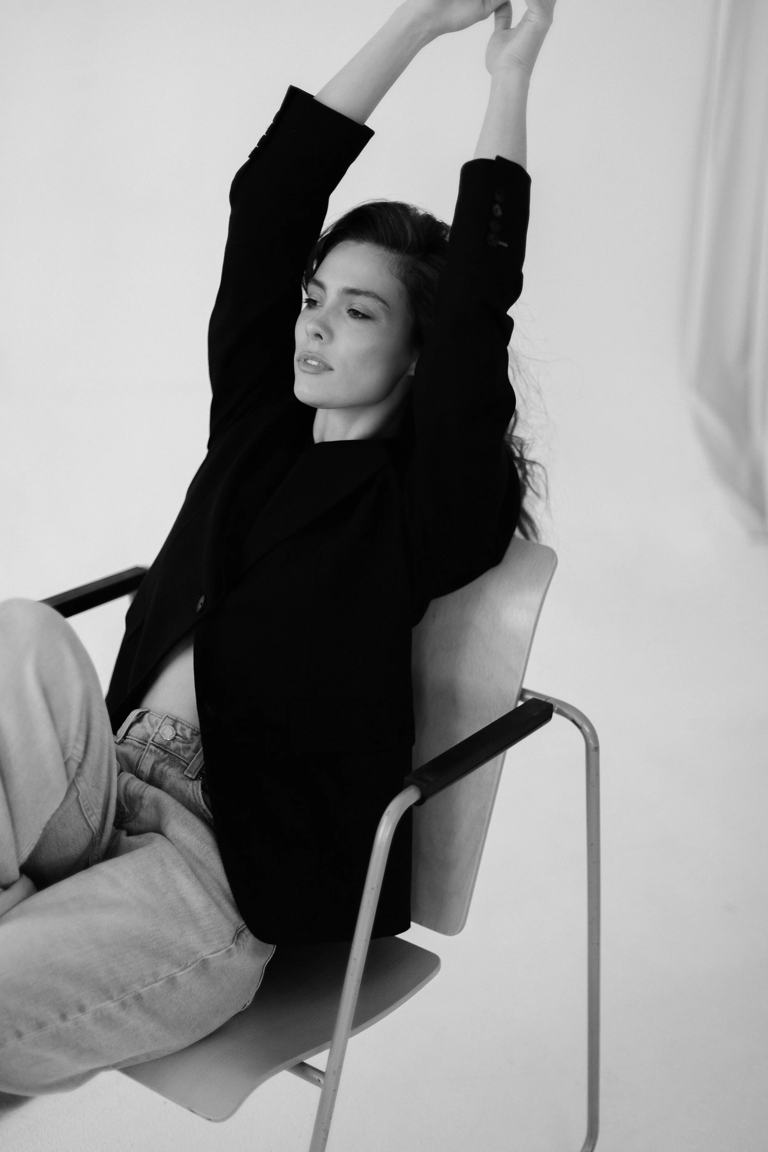 A woman with dark hair in a black blazer and light-colored pants sitting in a chair with her eyes closed, stretching her arms above her head.