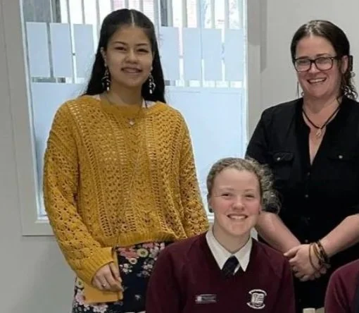 Three women smiling indoors in front of a window, two standing and one sitting, with casual and school attire.