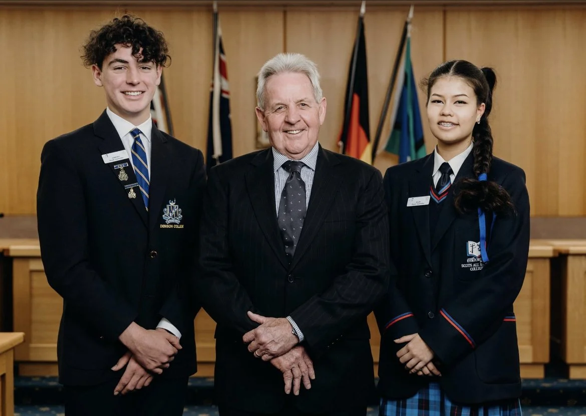 Three people, two students in uniforms and one older man in a suit, standing in a room with flags in the background.