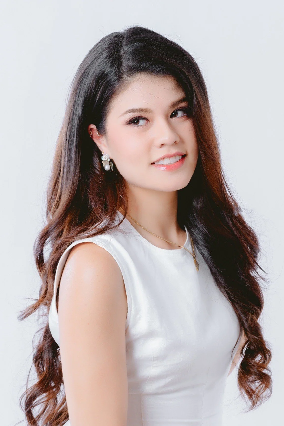 Portrait of a young woman with long wavy hair, wearing pearl earrings, a gold necklace, and a white sleeveless top, smiling slightly and looking to the side against a plain white background.
