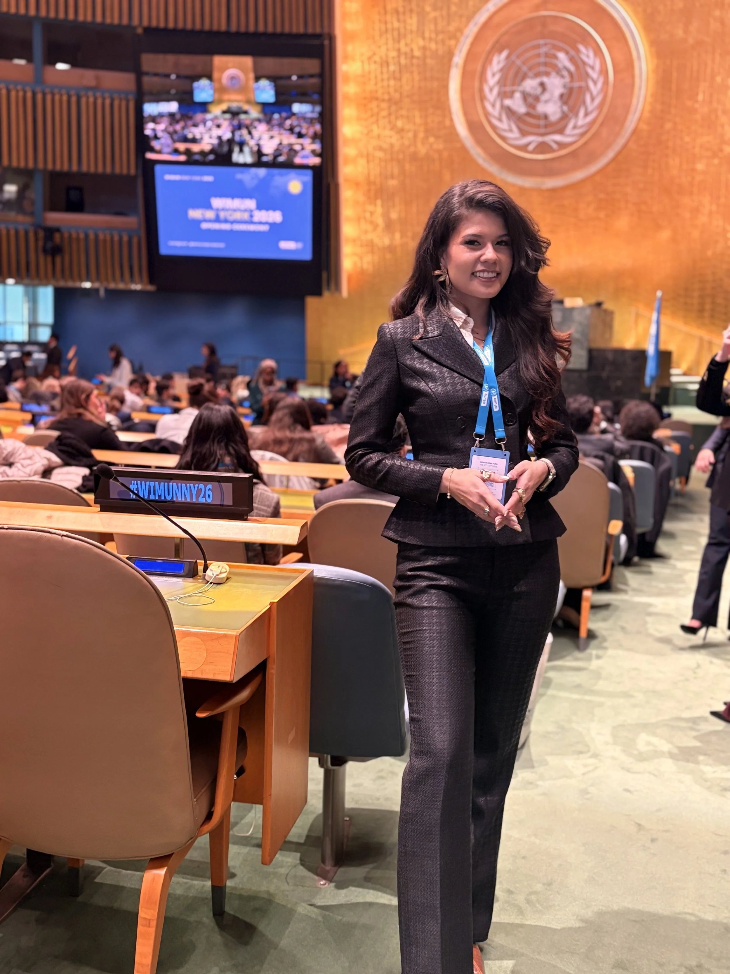 A woman with long dark hair dressed in a dark blazer and matching pants standing inside the United Nations meeting hall. The Women’s UN banner is on a desk in the foreground, and the UN emblem is on the wall behind her. The hall is filled with attendees.