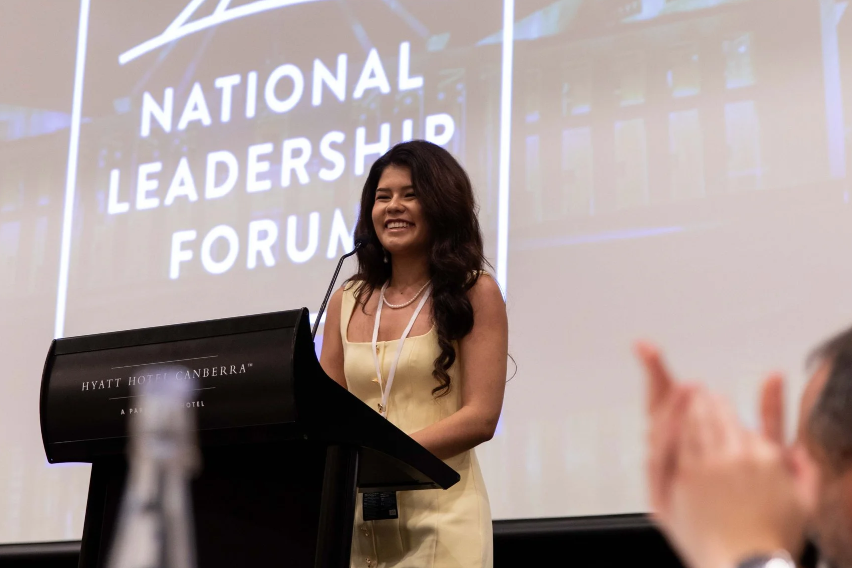 A woman with long dark hair, wearing a pale yellow dress and pearl necklace, stands at a podium with a microphone and smiles. Behind her is a large screen displaying the words 'National Leadership Forum'. The event is held at Hyatt Hotel Canberra.