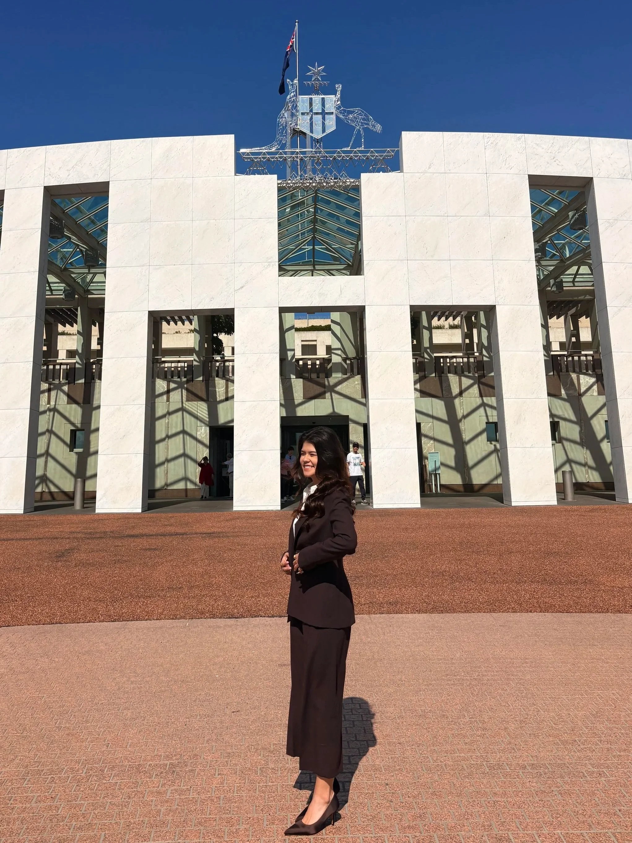A woman in a black suit and black heels stands smiling outside a modern building with white walls and large rectangular openings, with a blue sky overhead and an Australian flag on top of the building.