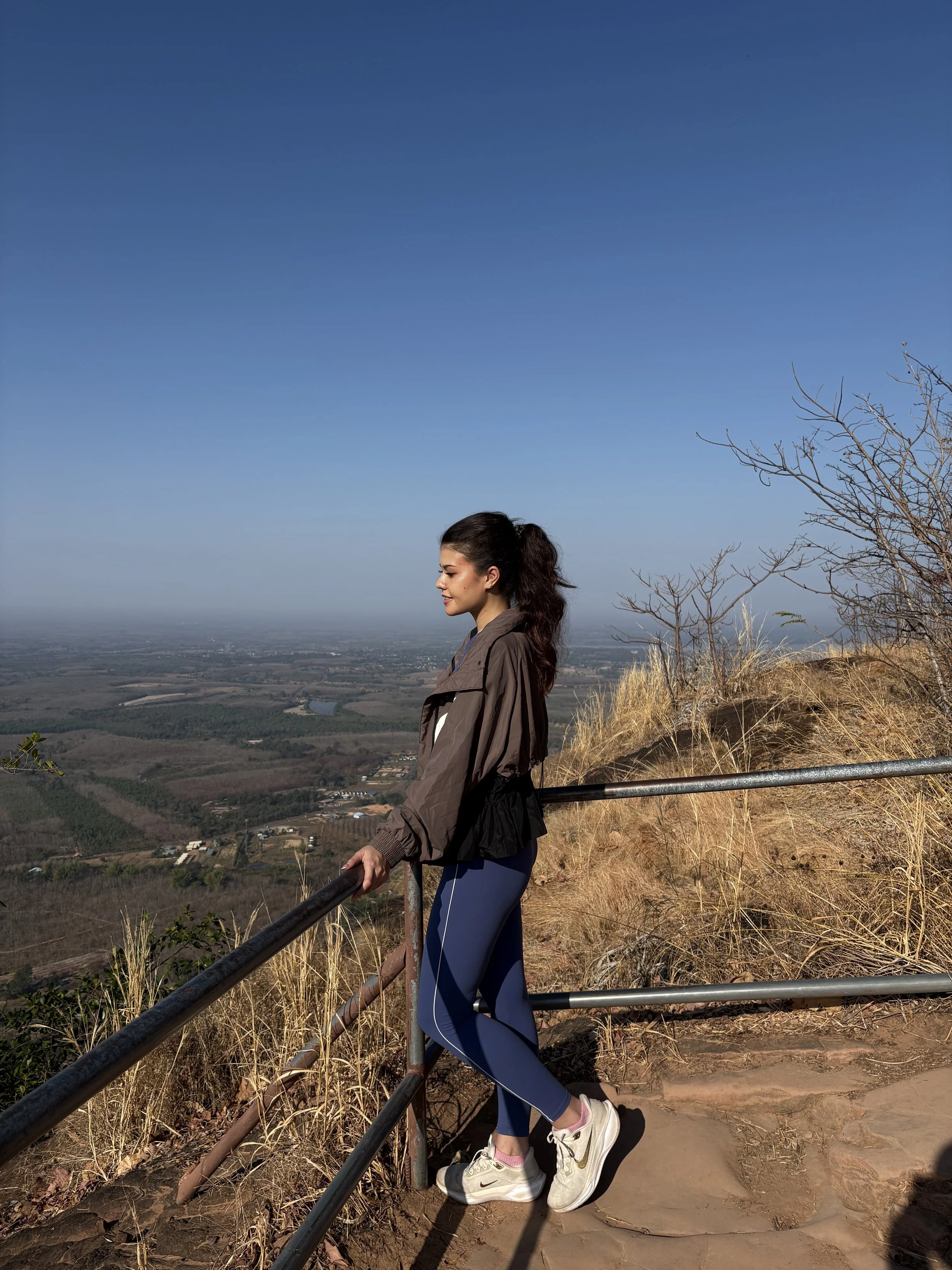 Young woman with long dark hair, tied back, standing on a hiking trail with a scenic view of valleys and rivers in the background, wearing a brown jacket, blue leggings, white sneakers, and looking downward.