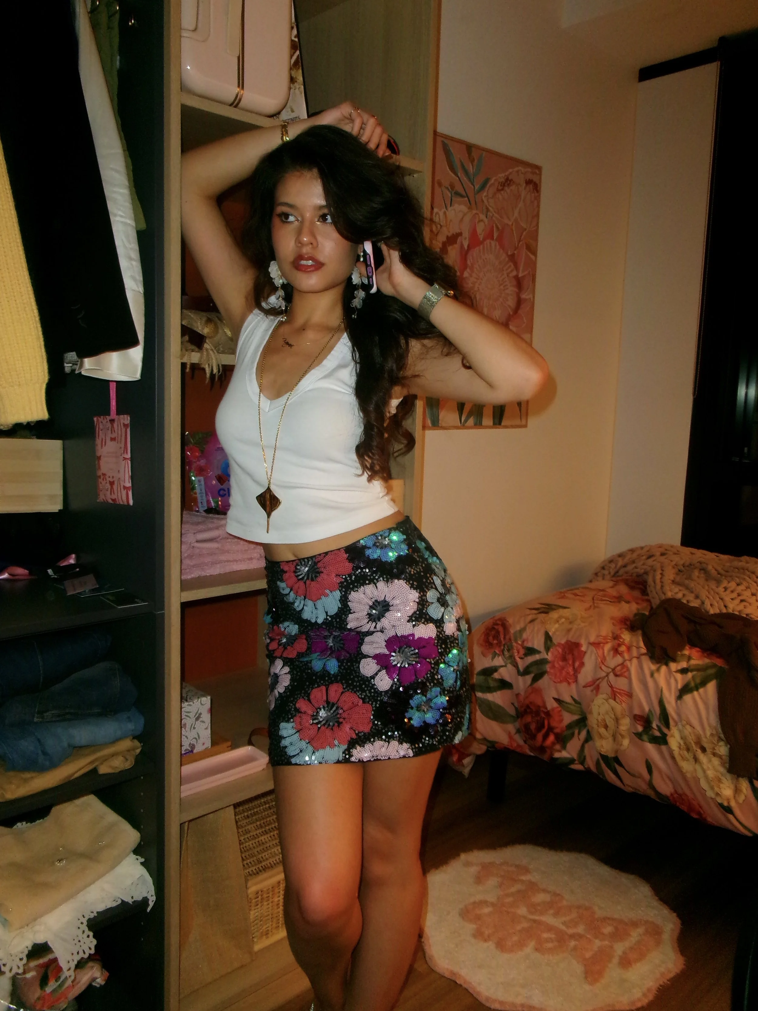 Woman with long dark hair wearing a white sleeveless top, colorful floral skirt, large earrings, and a necklace, standing in a bedroom in front of a mirror, with a bed and wardrobe in the background.