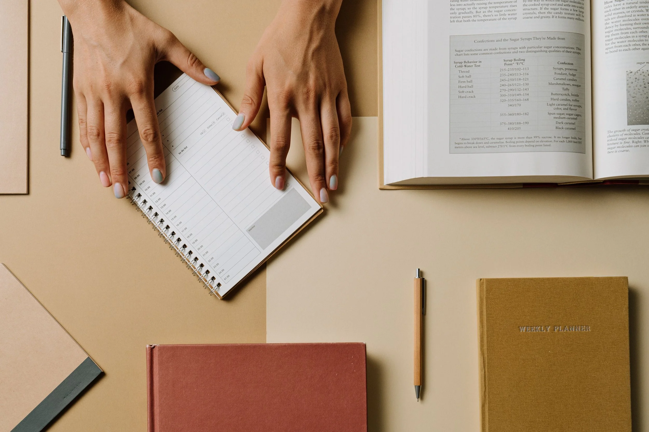 A person’s hands with light blue nails resting on an open spiral planner with a weekly schedule. The planner is on a beige table alongside a black pen, a closed red notebook, an orange pen, and another closed gold-colored planner labeled 'WEEKLY PLANNER.'